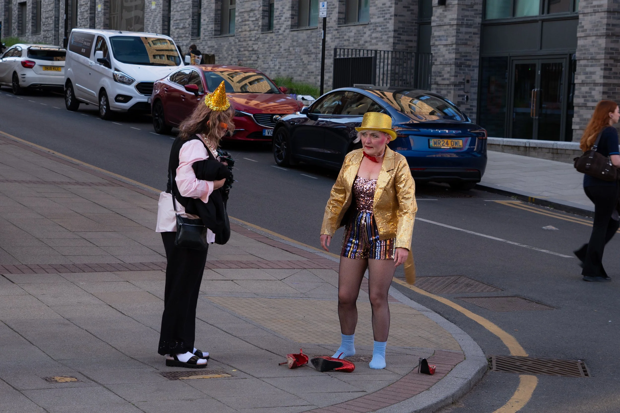 Two women dressed in colorful party costumes standing on a city sidewalk near parked cars. One woman wears a gold hat, sequin jacket, and fishnet stockings; the other has a birthday hat and casual overcoat.