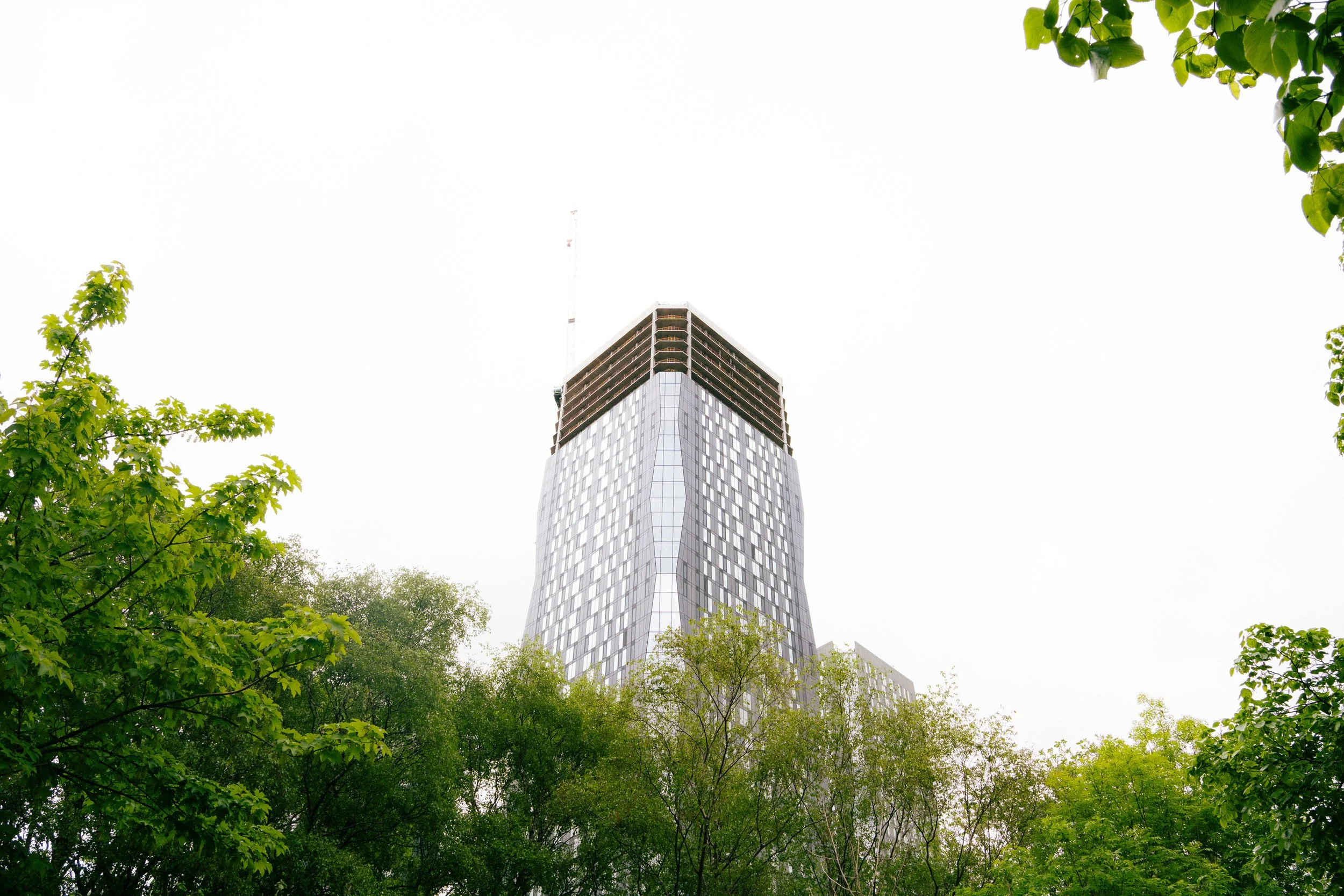 A tall modern skyscraper with glass windows is seen from below, surrounded by green leafy trees.