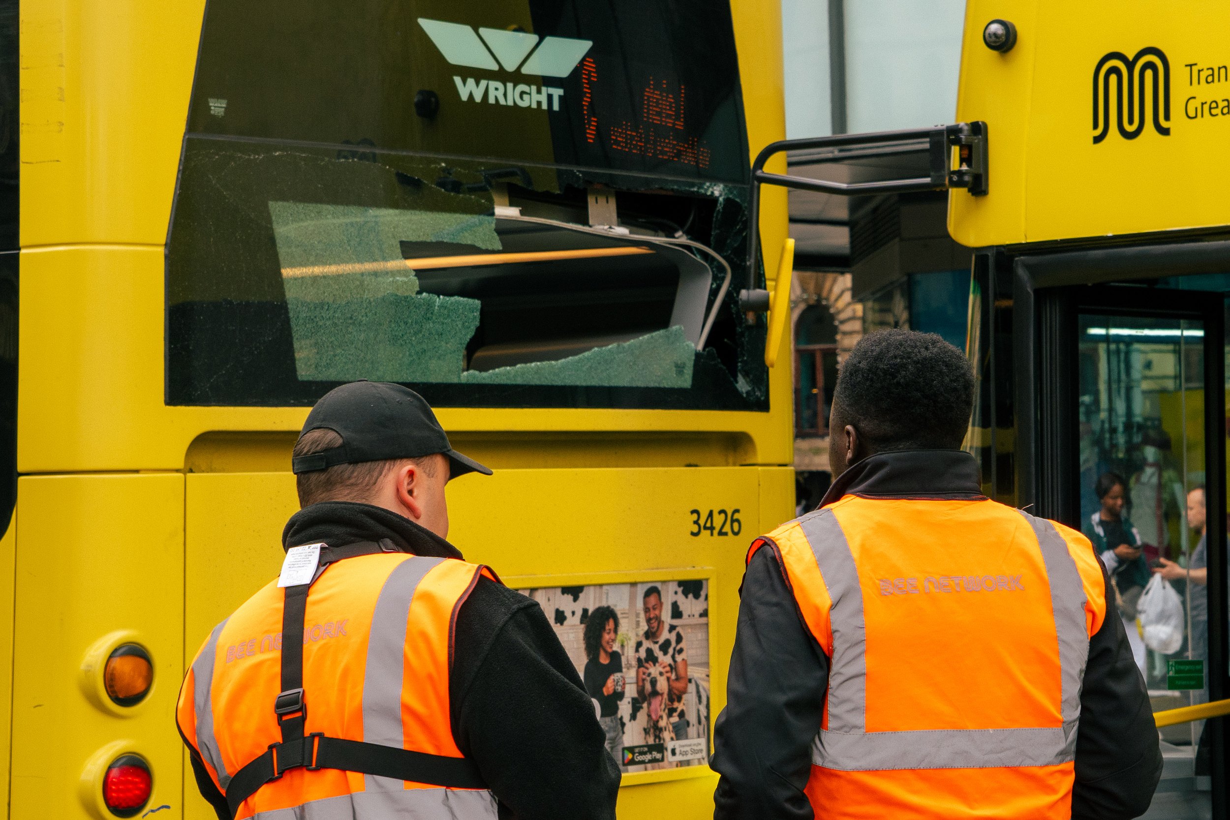 Two men with black hair wearing orange safety vests in front of a yellow bus with people visible in the background.