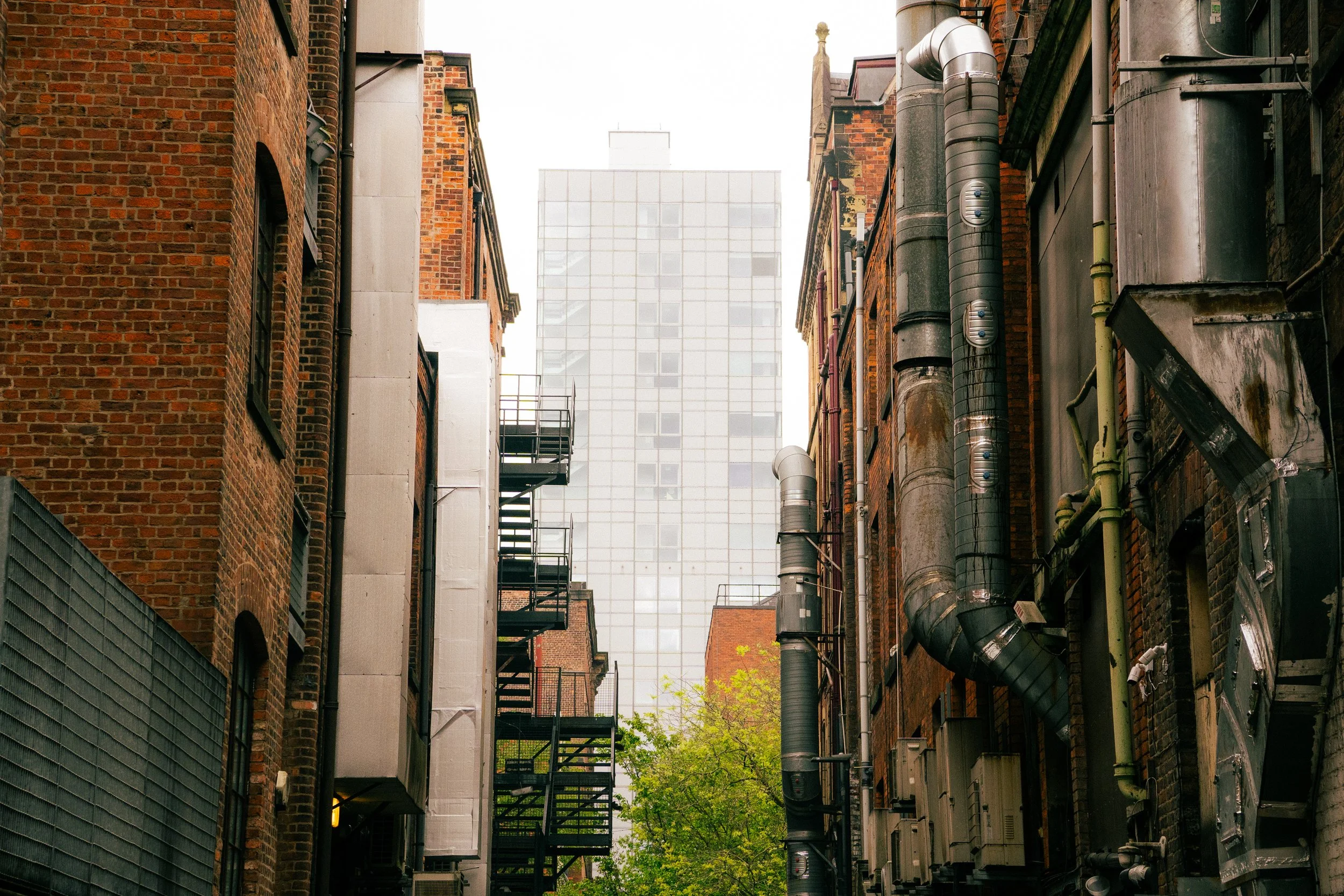 Narrow alleyway between brick buildings with fire escapes and various pipes, with a modern glass building in the background.
