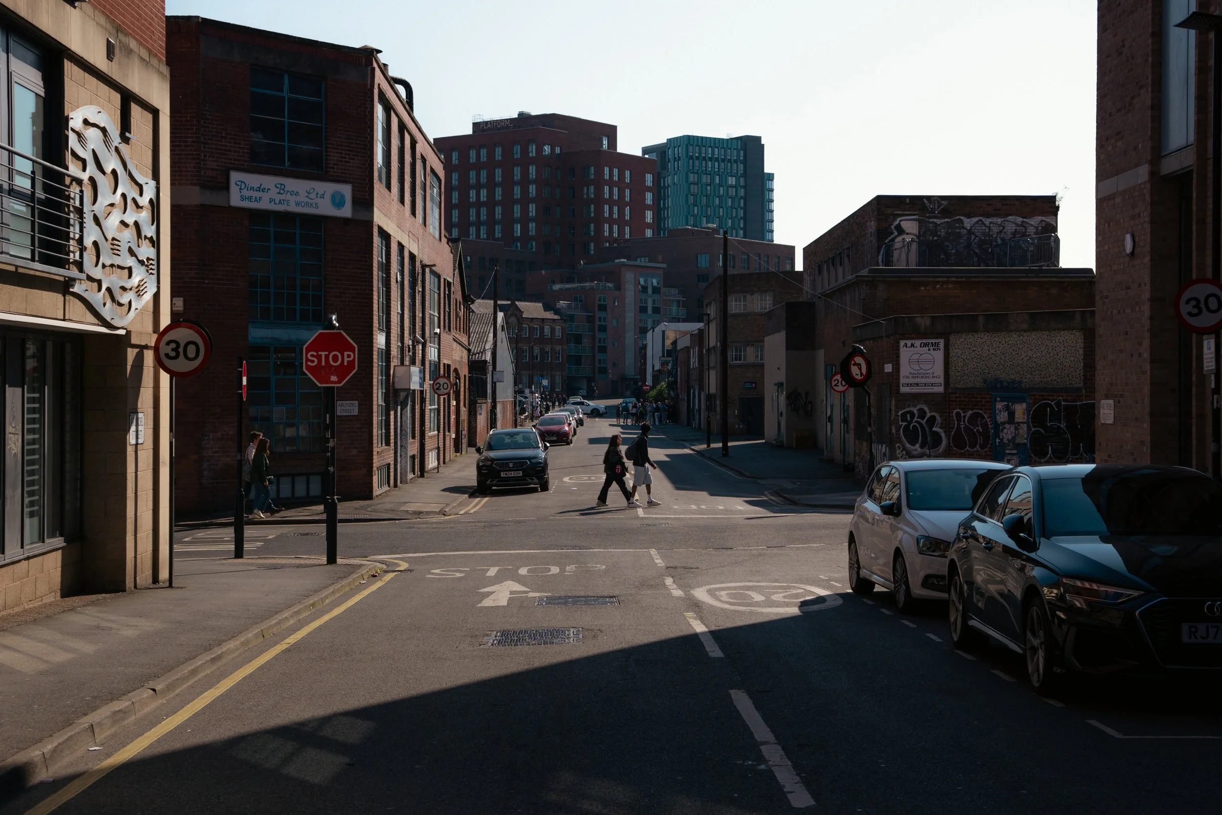 A city street with parked cars and pedestrians walking on the crosswalk, surrounded by brick and modern buildings under a clear sky.