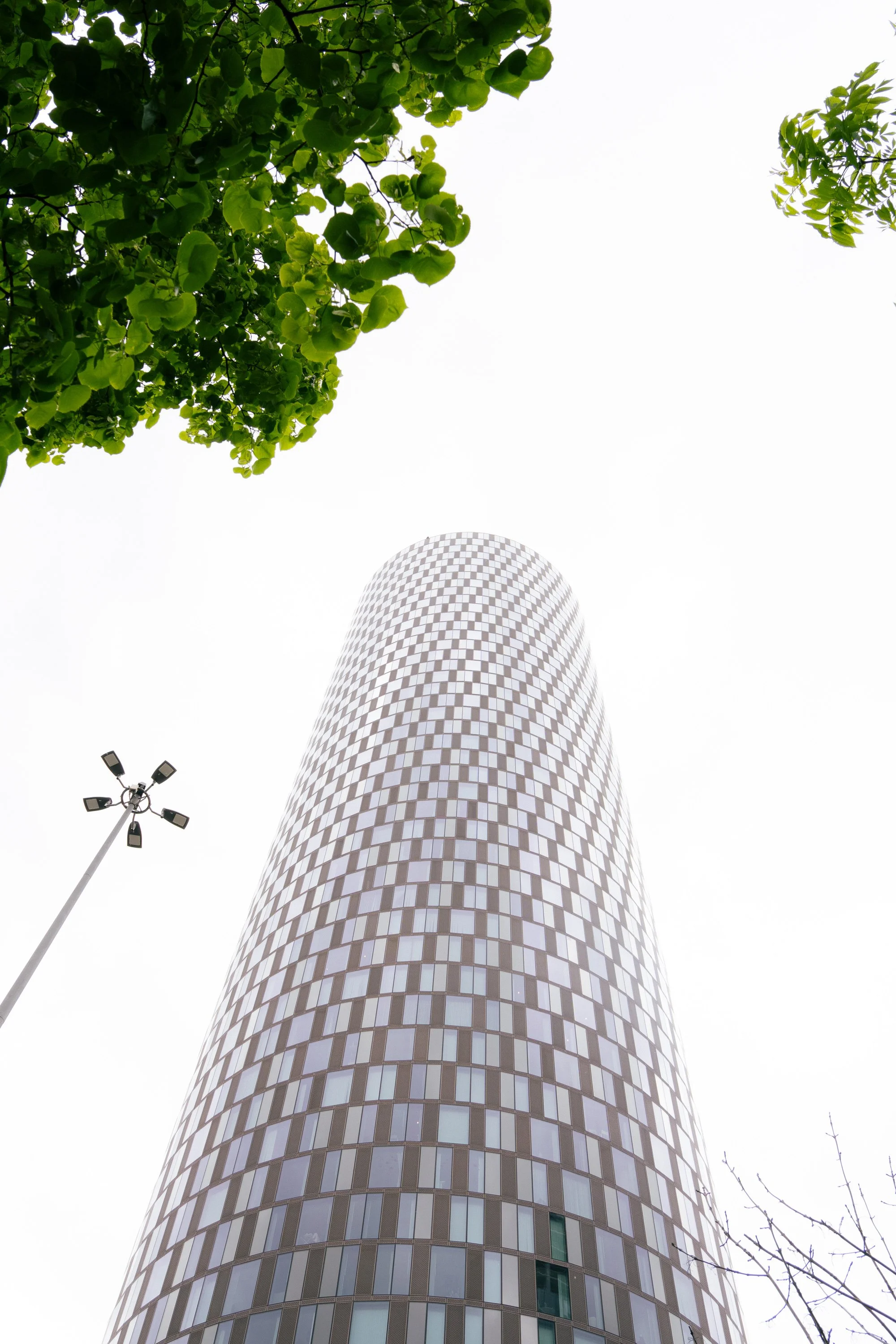 Looking up at a tall, cylindrical building with a checkered pattern on its facade, with green tree leaves on the top left corner and a cloudy sky background.