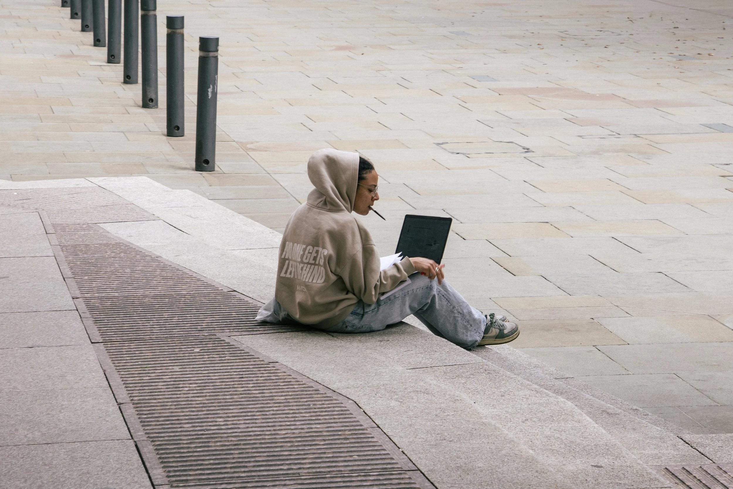 A young woman with glasses, wearing a beige hoodie and gray sweatpants, sitting on the sidewalk with a laptop and papers, smoking a cigarette.