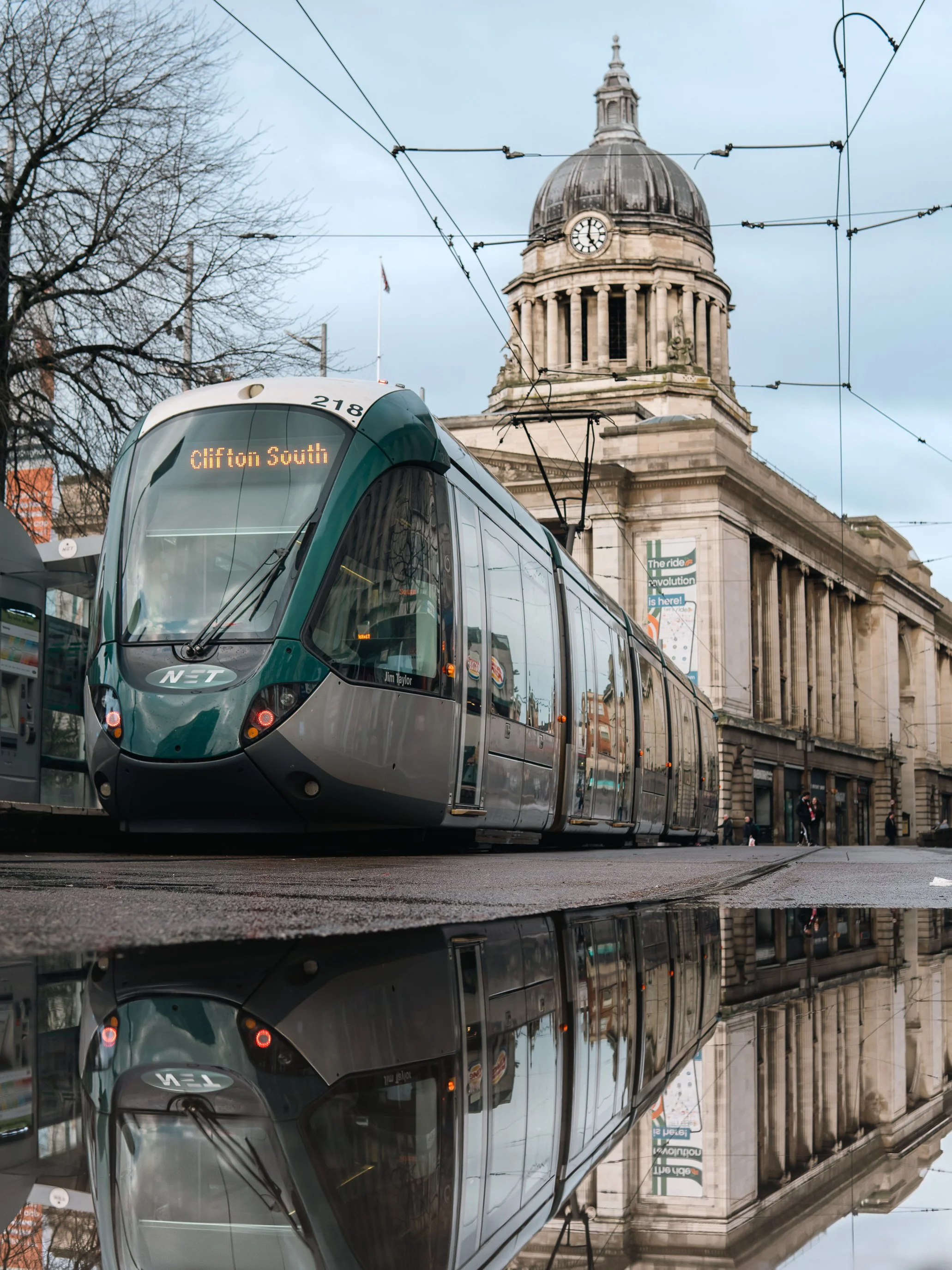 A modern tram with the sign 'Clifton South' on the front, traveling on tracks in front of a historic building with a clock and a dome, and some leafless trees in the background.