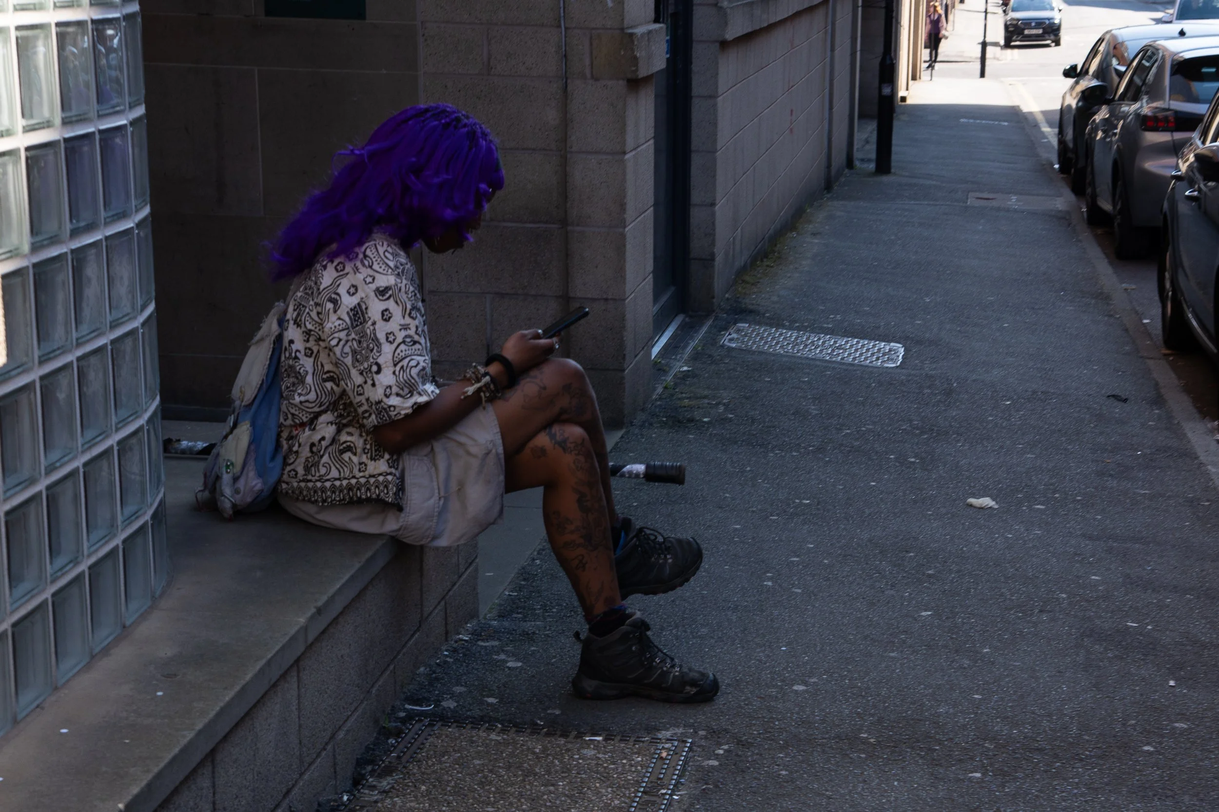 A person with vibrant purple hair sitting on a sidewalk, looking at their phone, beside a brick building with glass blocks, in an urban street scene with parked cars.