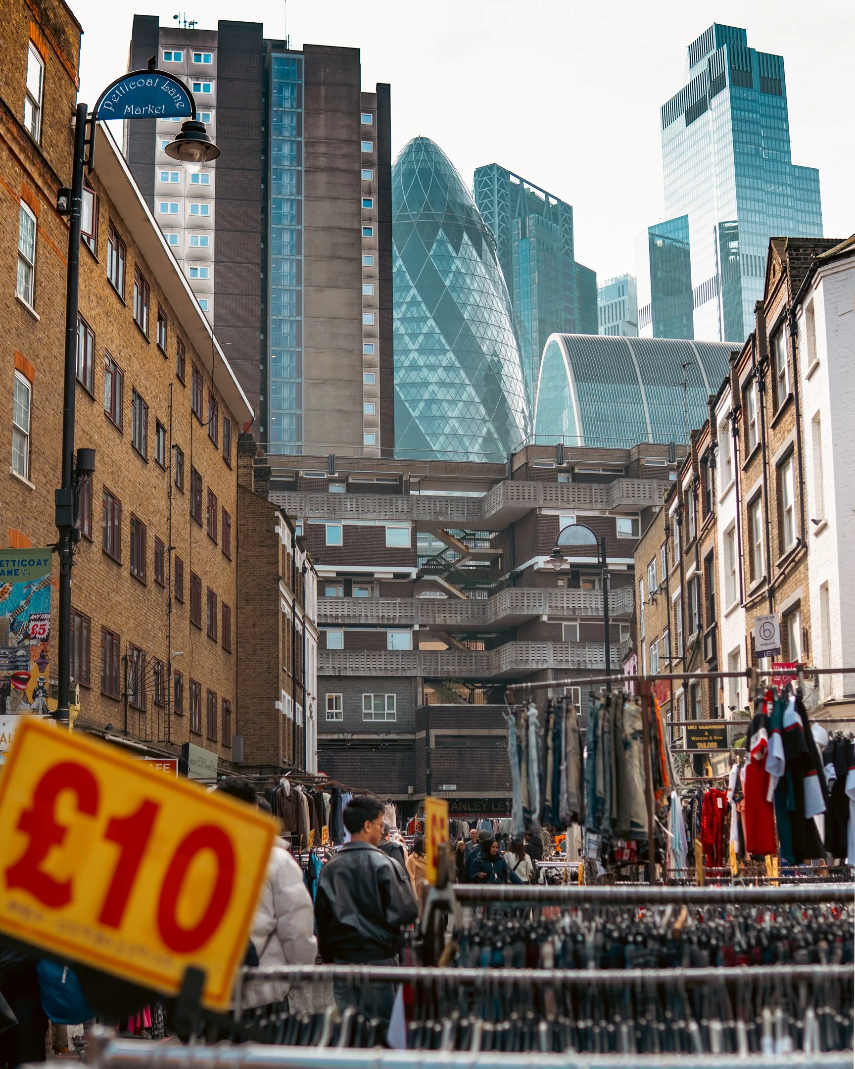 Street market with clothing stalls, people shopping, and tall modern skyscrapers in the background.
