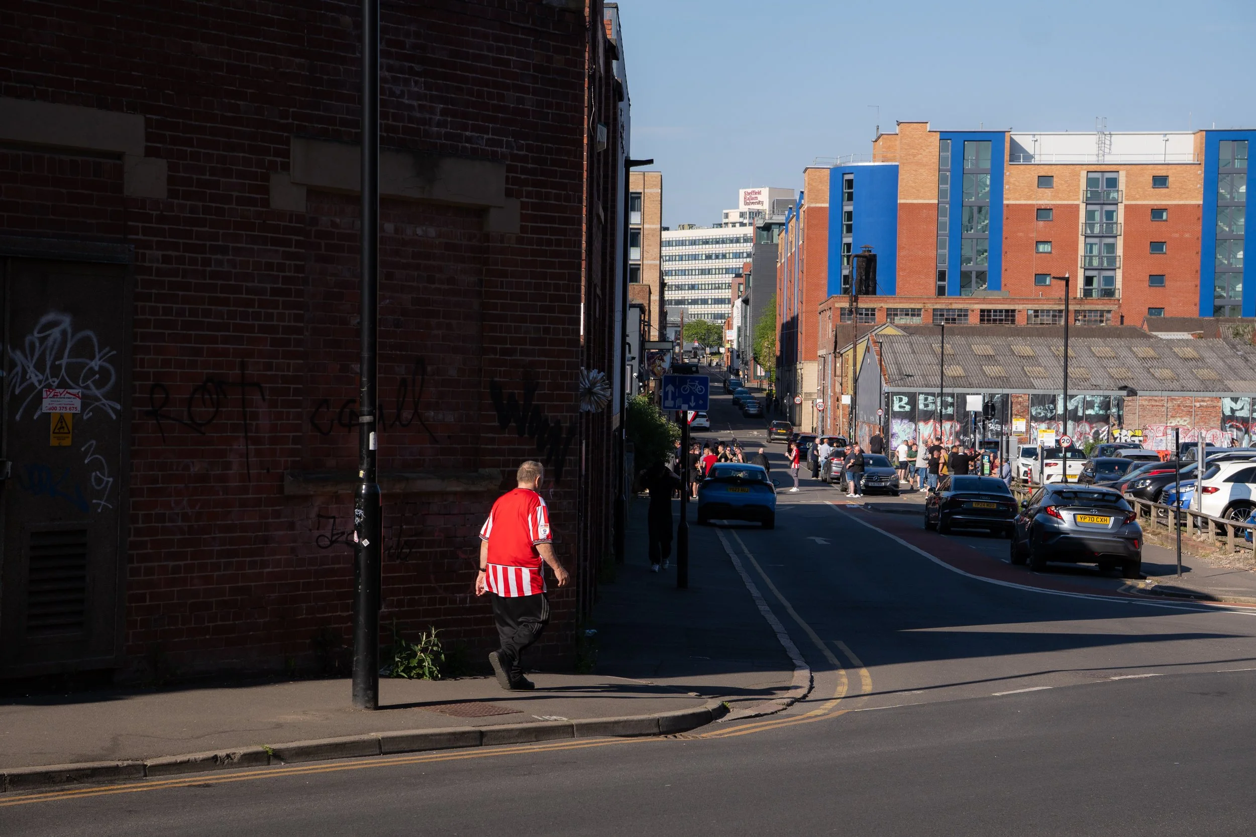 City street scene with modern and older buildings, cars parked along the road, and pedestrians walking. A man in a red sports jersey and striped shorts walks on the sidewalk in the foreground. The sky is clear and blue.