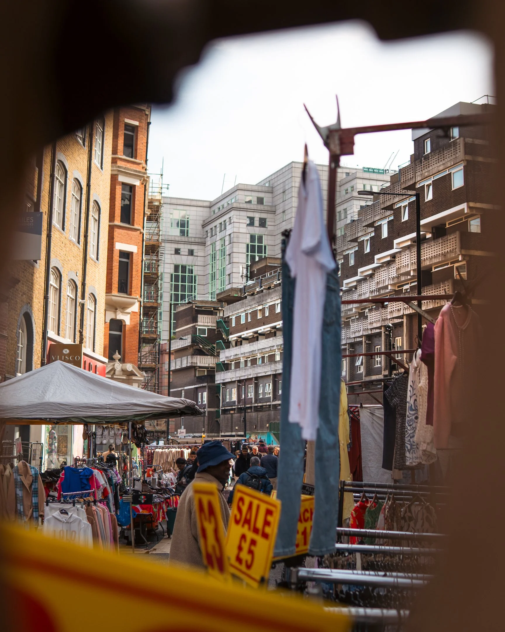 Street market with clothing stalls, sale signs, and people shopping amid tall buildings in an urban area.