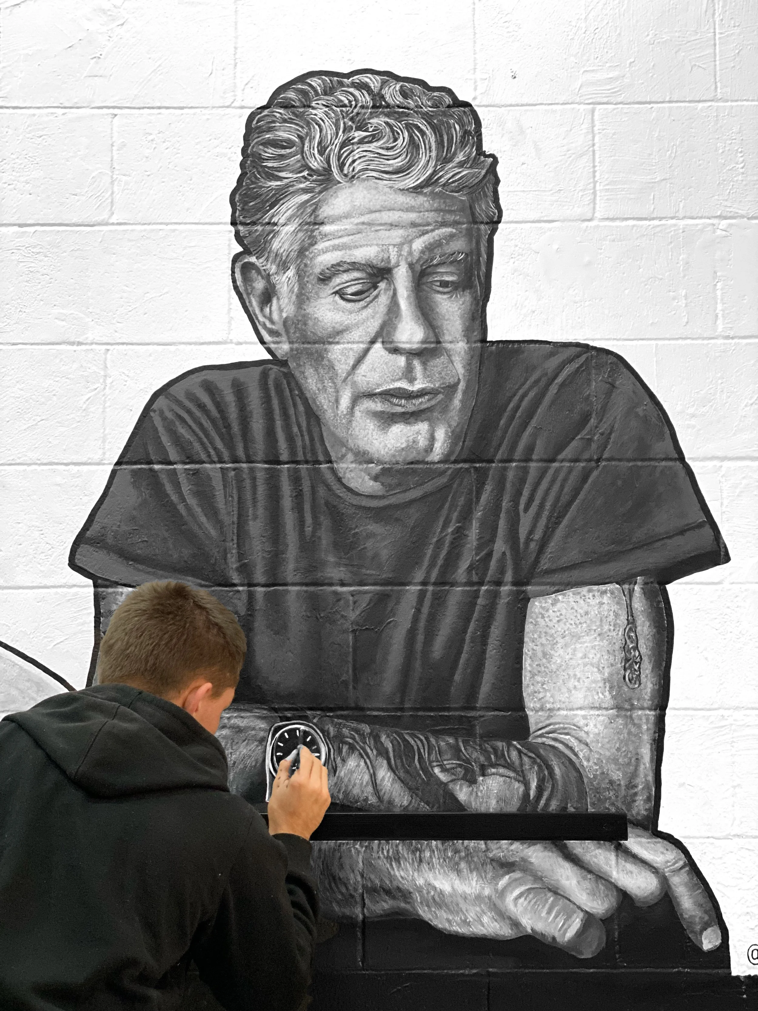 A person drawing a black and white mural of a man with wavy hair and closed eyes on a brick wall.