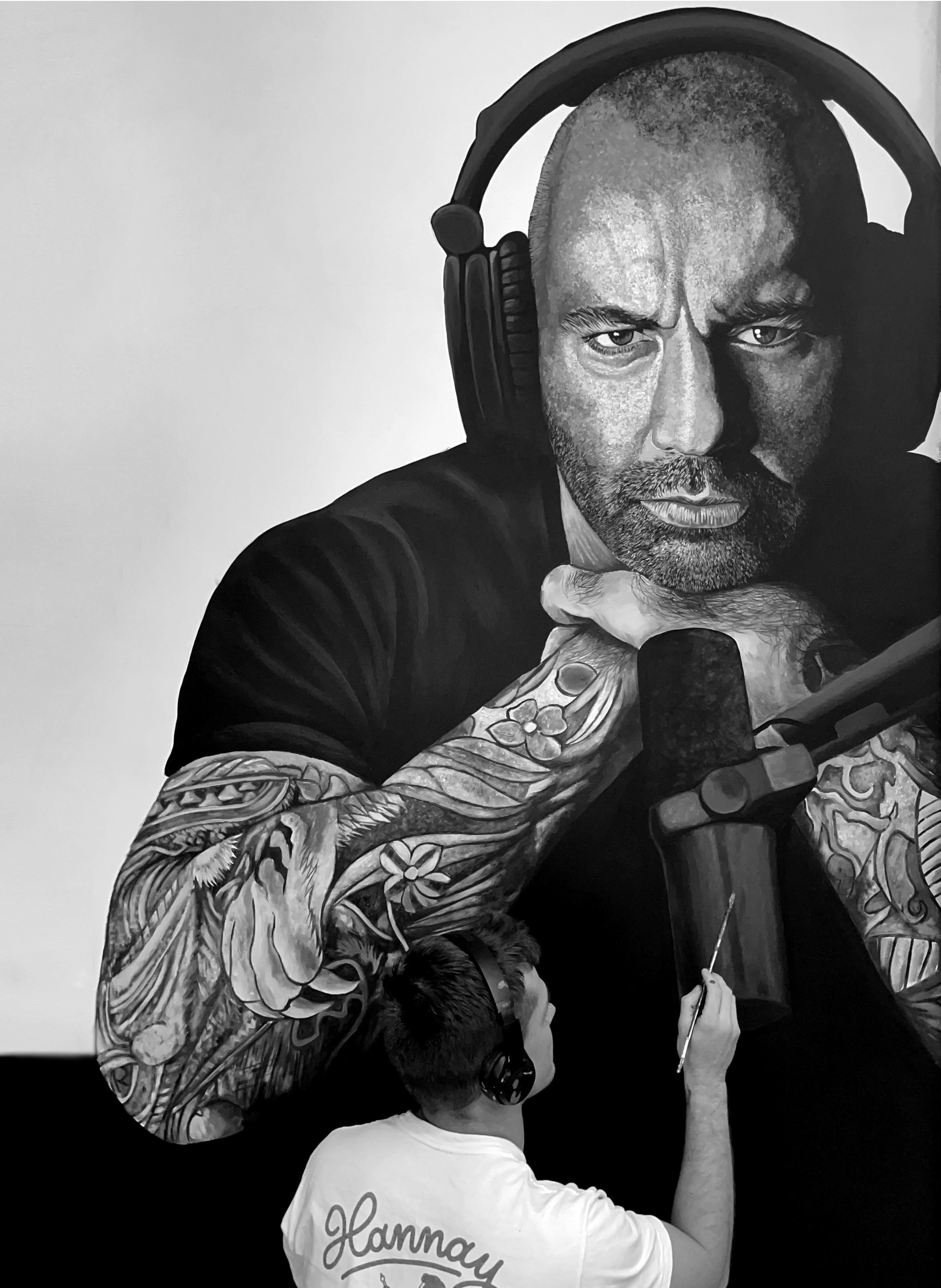 Black and white mural of a man with a shaved head and tattoos on his arm, wearing headphones and holding a microphone, with a person in a shirt labeled 'Hannah' taking a photo in front of it.