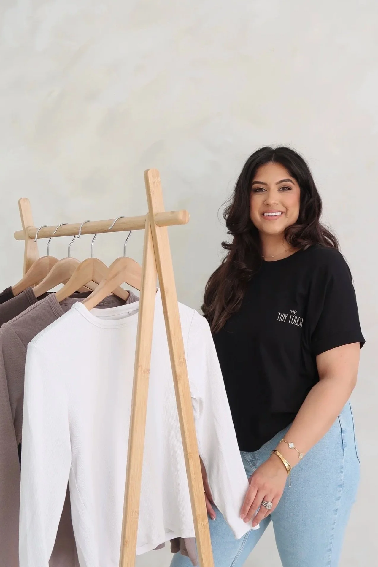 A woman with dark hair smiling, standing next to a clothing rack with white and brown tops on wooden hangers.