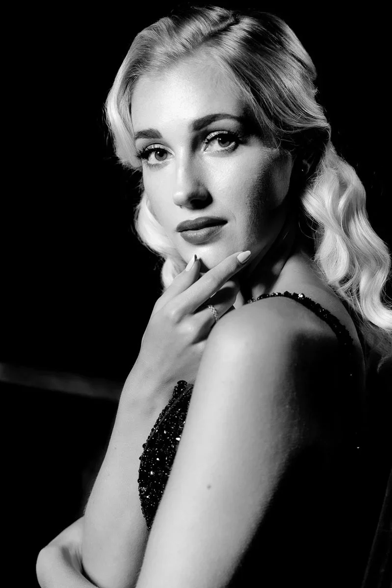 Black and white portrait of a woman with wavy blonde hair, wearing a sparkly dress, with her hand near her face, looking at the camera.