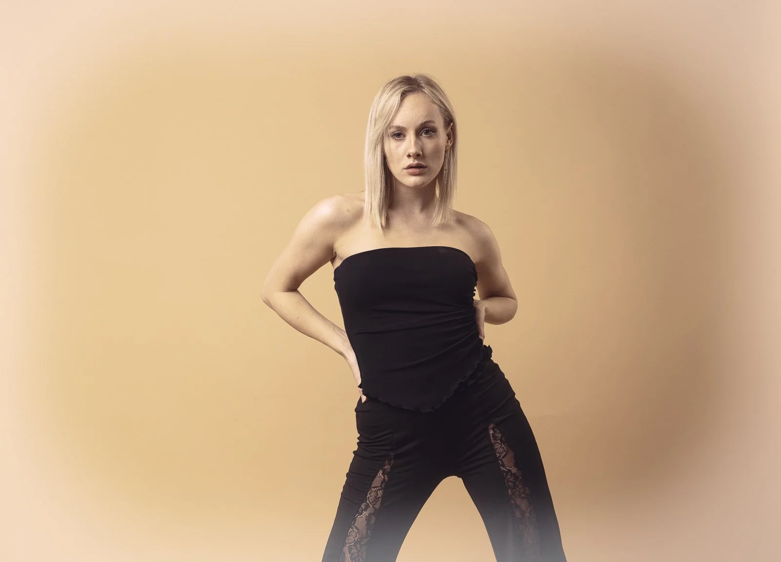 A young woman with blonde hair, wearing a black strapless top and black pants with lace panels, posing against a beige background with her hands on her hips.