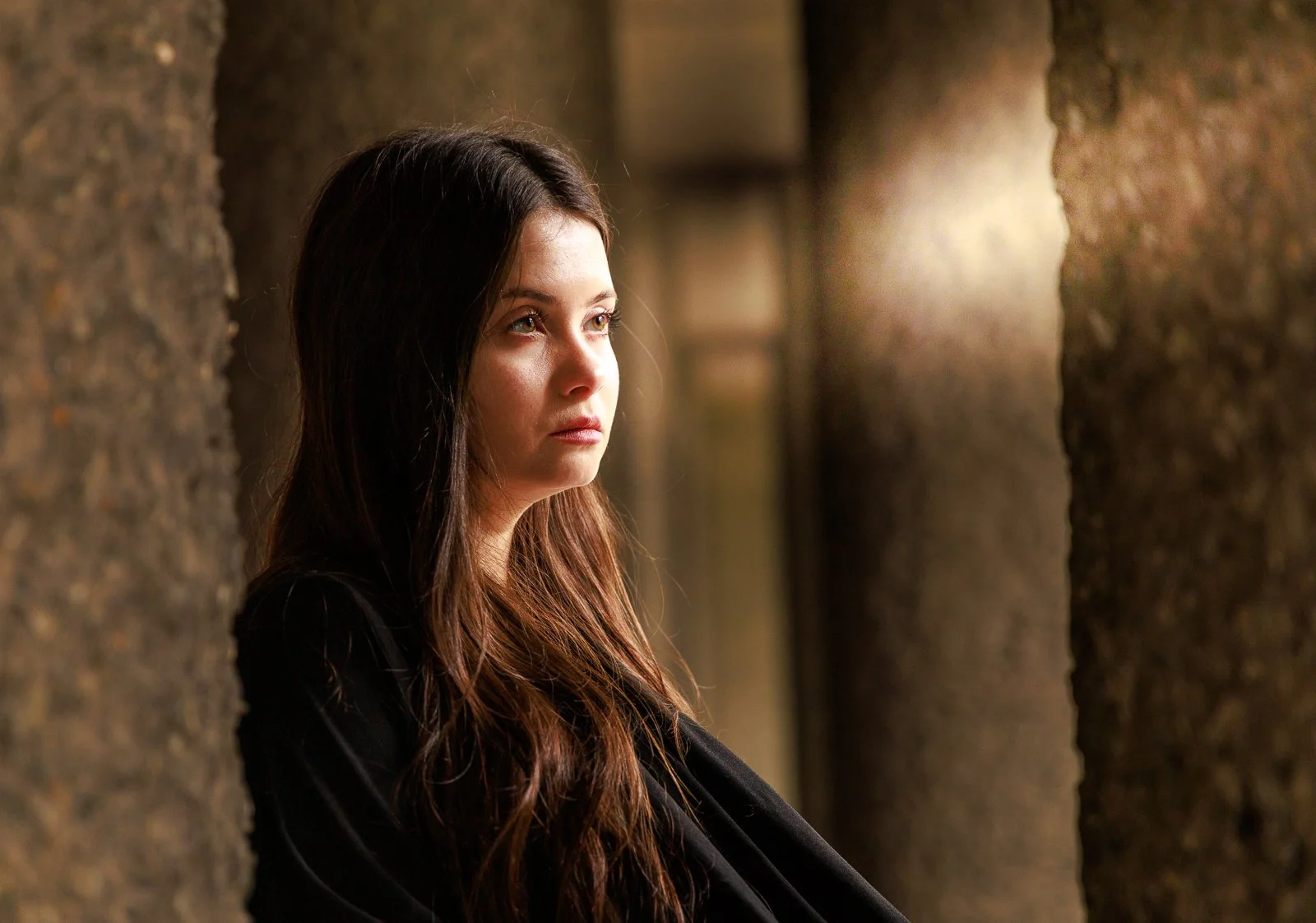 A young woman with long dark hair and light skin looking thoughtfully to the side, framed by rough-textured stone walls in a dimly lit setting.