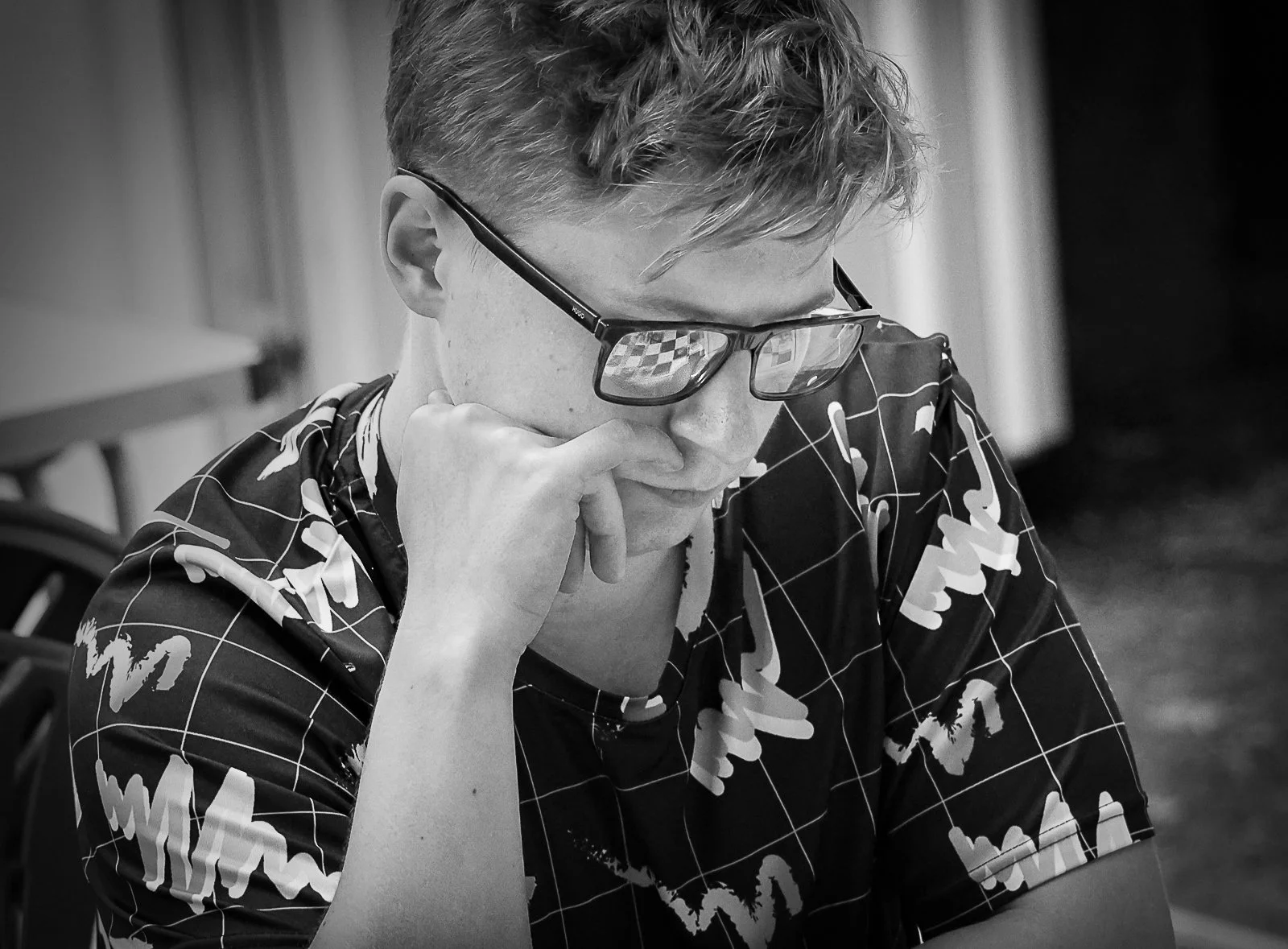 A young man with short hair and glasses sitting at a table, resting his head on his hand, looking down in thought, wearing a patterned shirt.