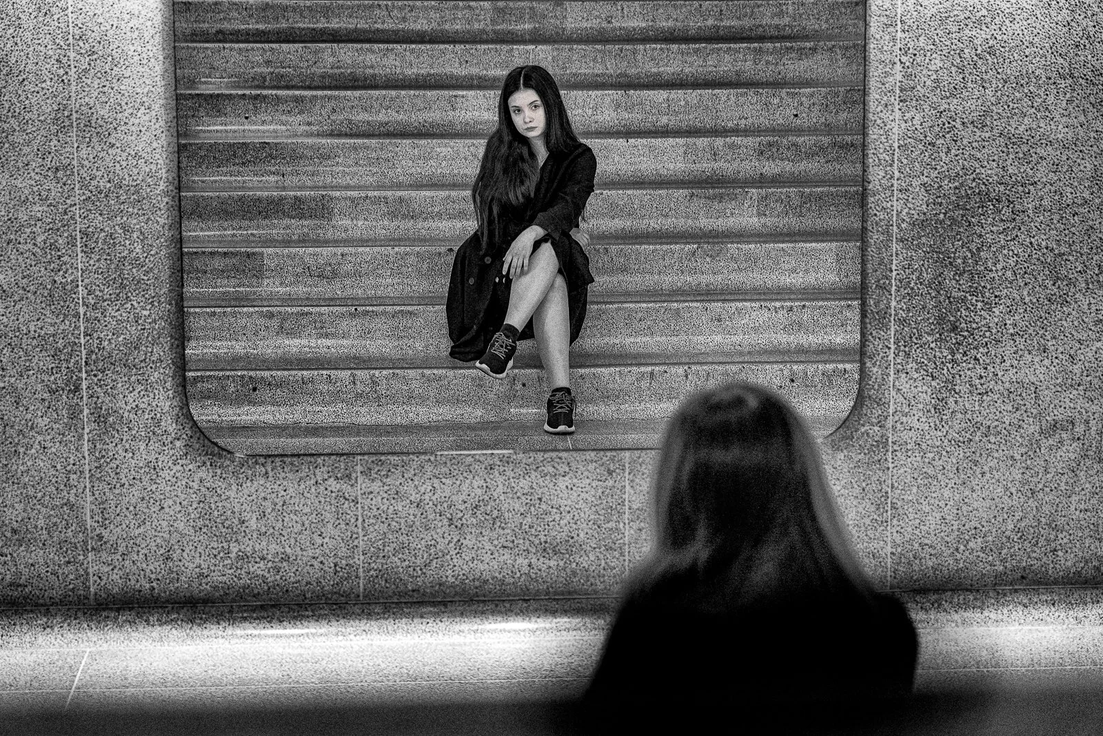 Black and white photo of a woman sitting on stairs in a framed window, with a person with long hair sitting on the floor in front of it, facing her.