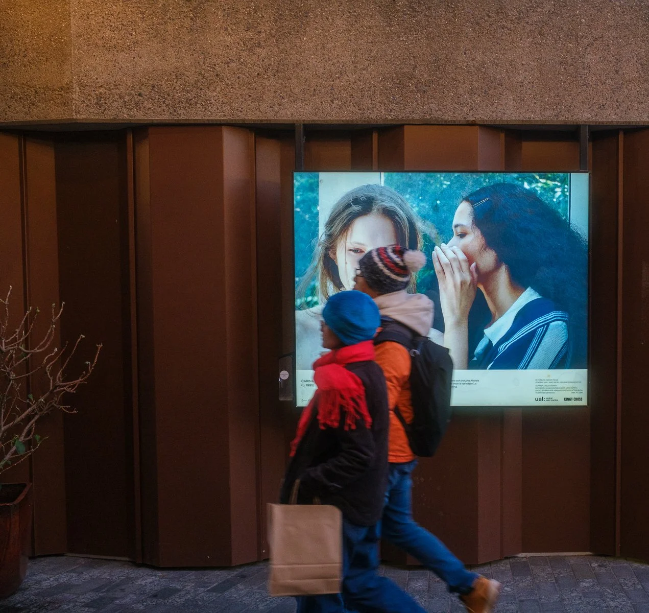 Two children walking past a digital advertisement in a building lobby. The advertisement features a close-up of two women whispering to each other outdoors.