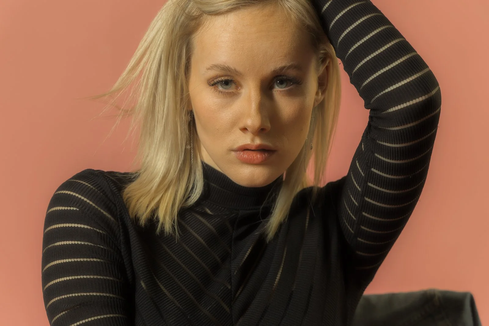 A young woman with blonde hair wearing a black top with sheer striped sleeves, posing with her left arm raised, against a pink background.