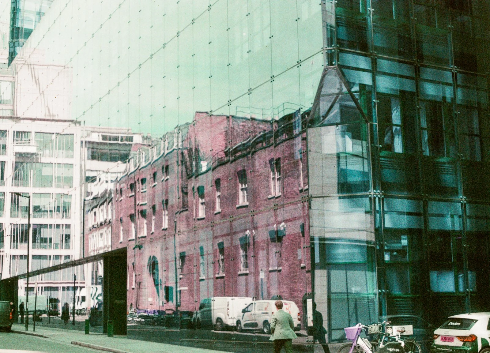 Reflection of historic brick building on modern glass building in an urban street scene, with parked cars, bicycles, and pedestrians.