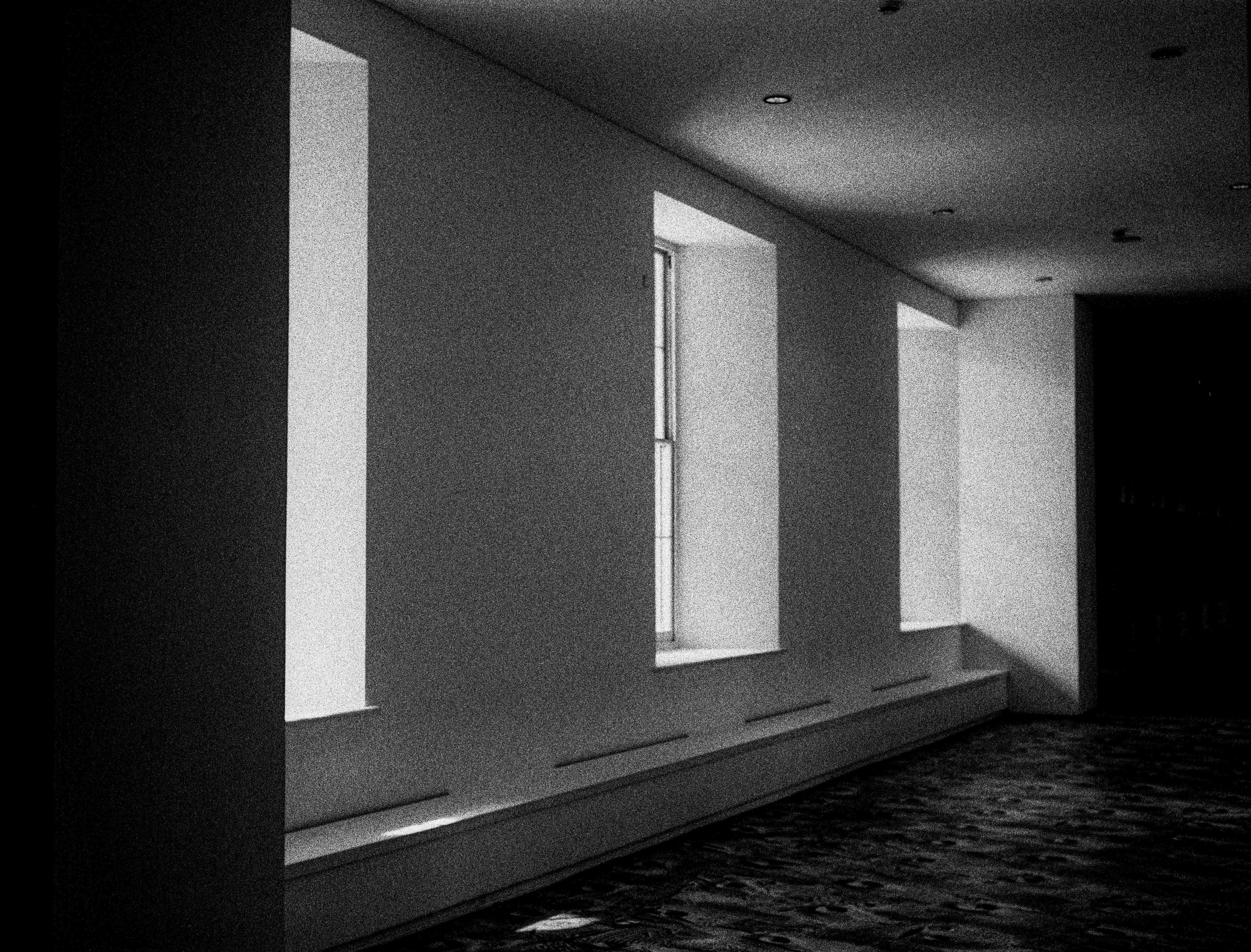 A black and white photo of an interior space with high windows, ceiling lights, and built-in furniture along the wall.