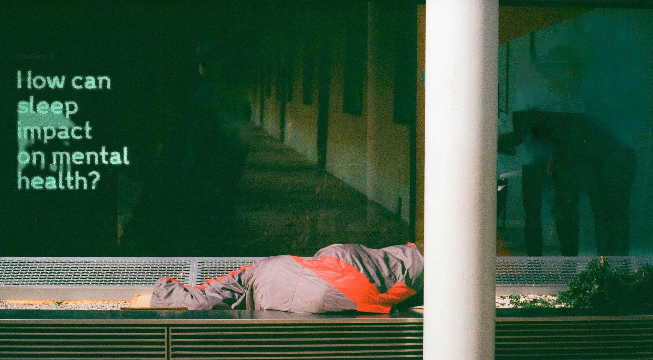 A person lying face down on the floor wrapped in a gray and orange sleeping bag in front of a dark green wall with white text about sleep and mental health.