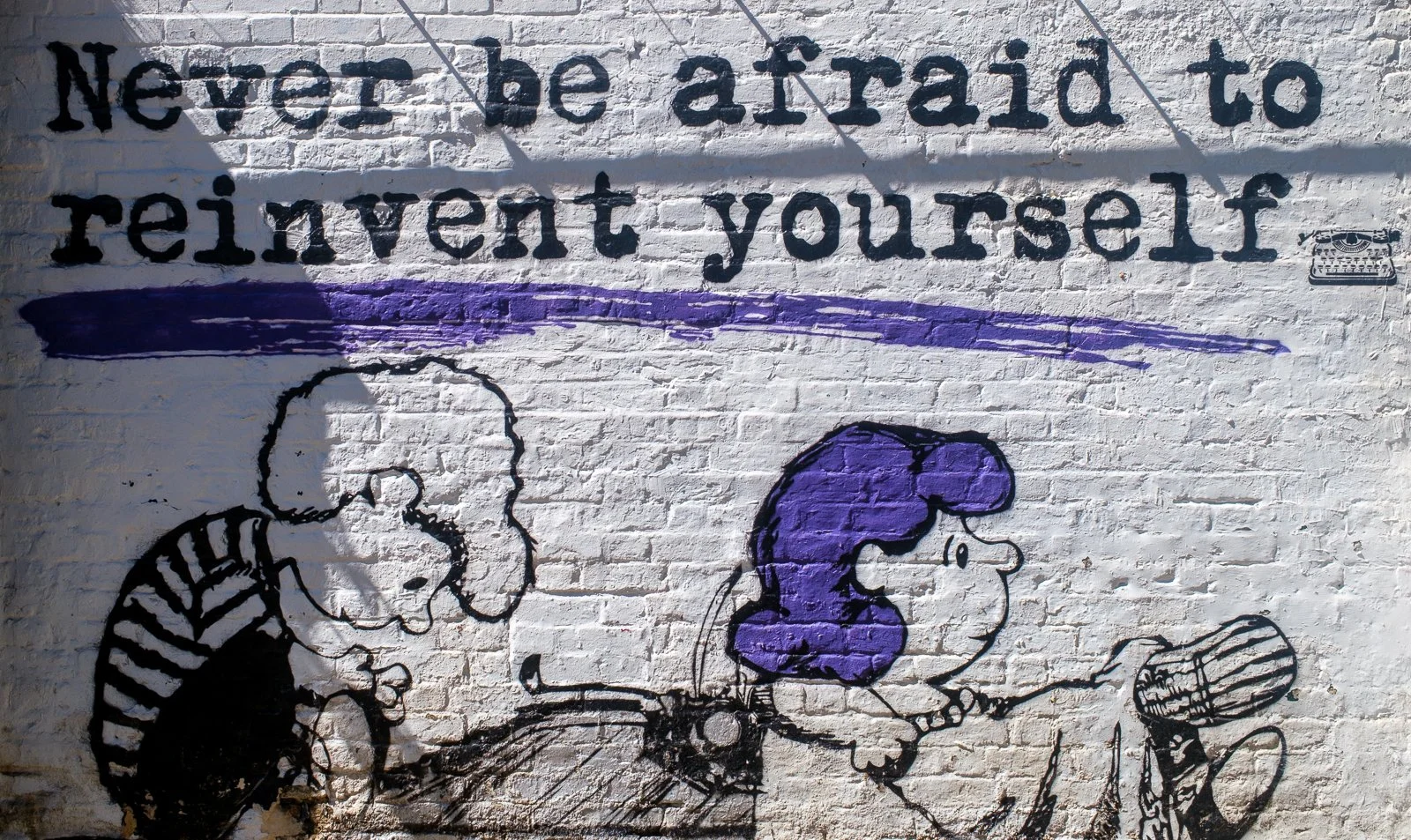 Graffiti mural on a white brick wall with the quote 'Never be afraid to reInvent yourself' in black letters, featuring a drawing of two children, a boy and a girl with purple hair, riding a bicycle, and a small decal of a typewriter in the top right corner.