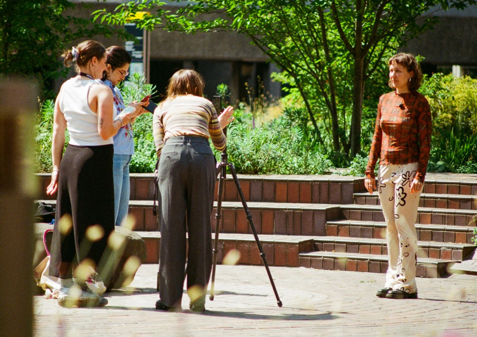 A woman is standing on steps outdoors in front of a green bush, being photographed by four women who are looking at their phones or camera. One of the women is adjusting her position, while the others are observing.