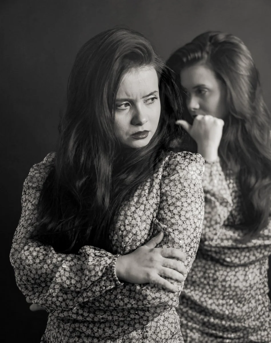 Black and white photo of two women with long hair, one in front with crossed arms looking annoyed and one in back whispering.