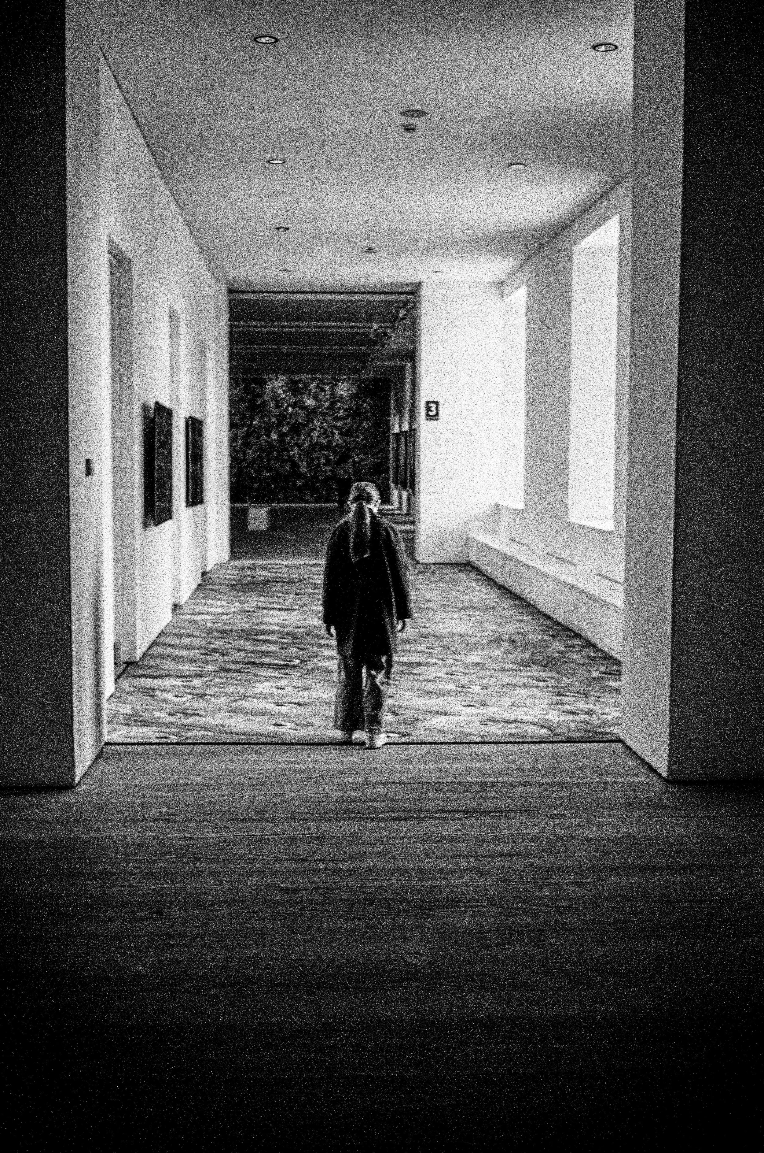 A person walking down a corridor with a patterned floor in a building, with artwork on the left wall and large windows on the right.