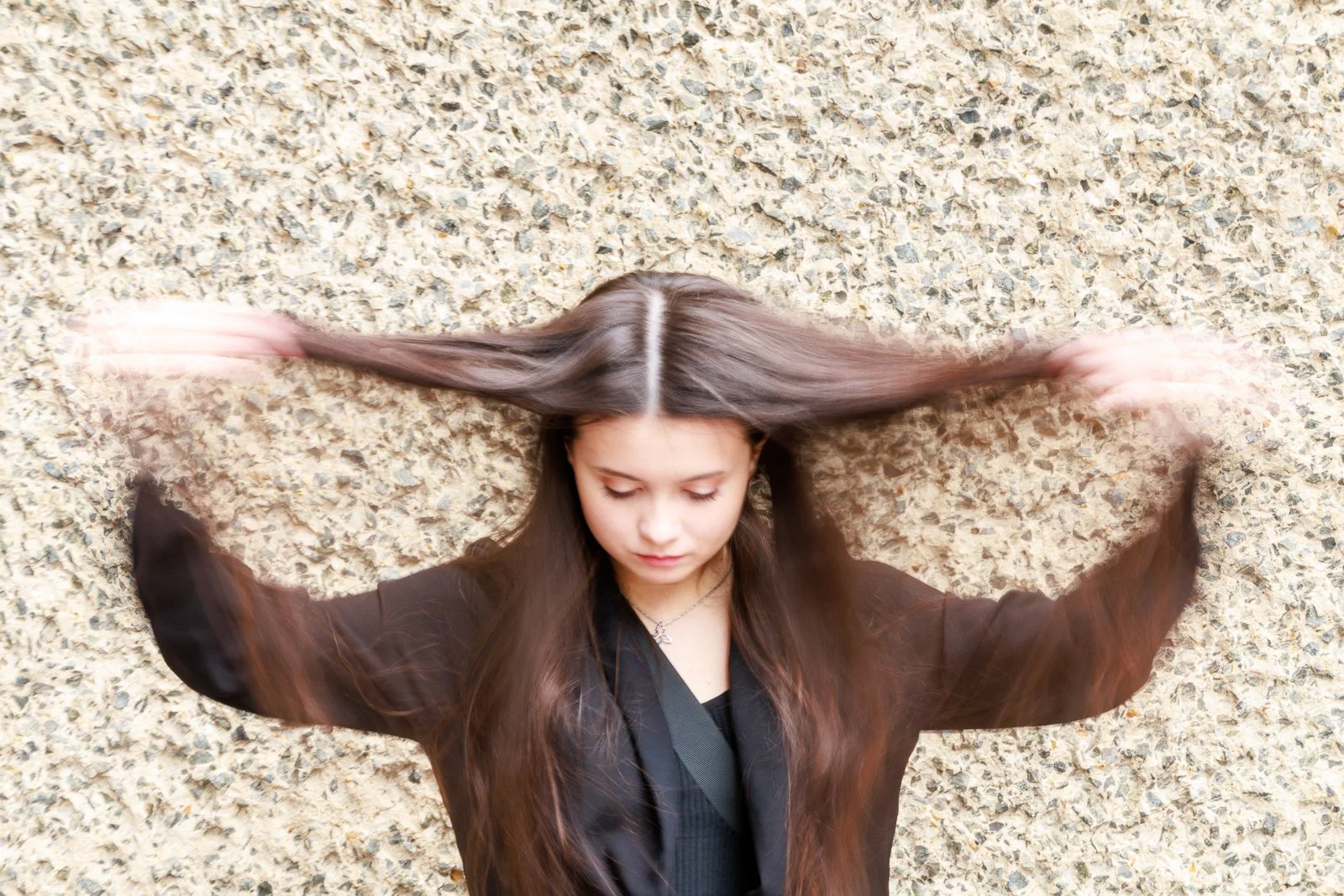 A young woman with long brown hair is lying on a textured beige wall, flipping her hair with her eyes closed and arms raised.