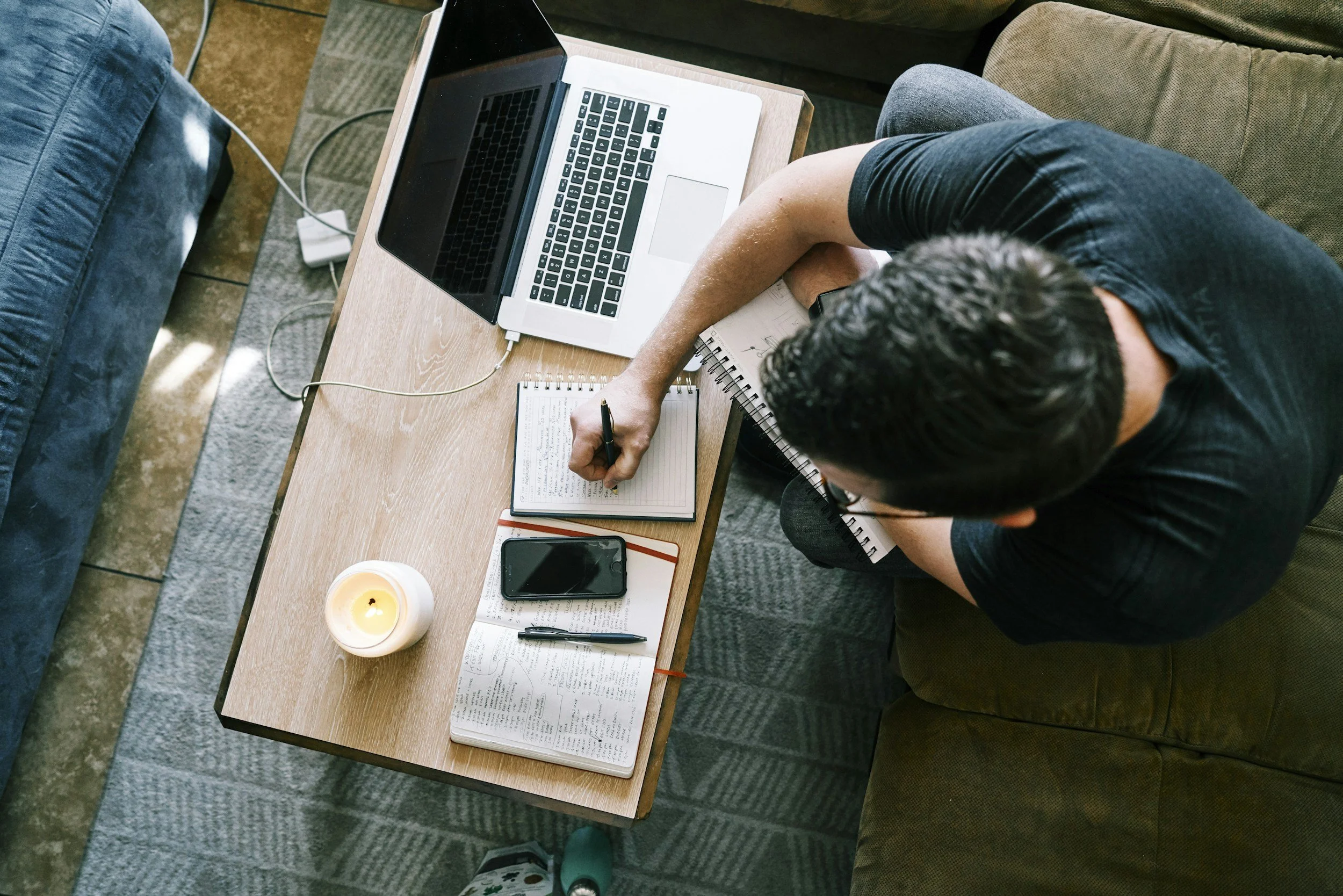 Overhead view of a person working at a wooden table with a laptop, notebook, pen, and smartphone, surrounded by a candle and a cup of tea or coffee. The person is writing in the notebook with a pen.