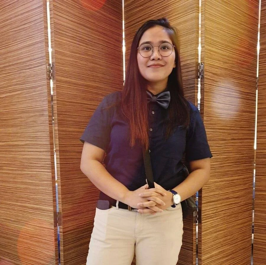 A young woman with glasses and straight hair, wearing a dark shirt with a bowtie, beige pants, and a wristwatch, standing in front of a wooden foldable partition, smiling at the camera.