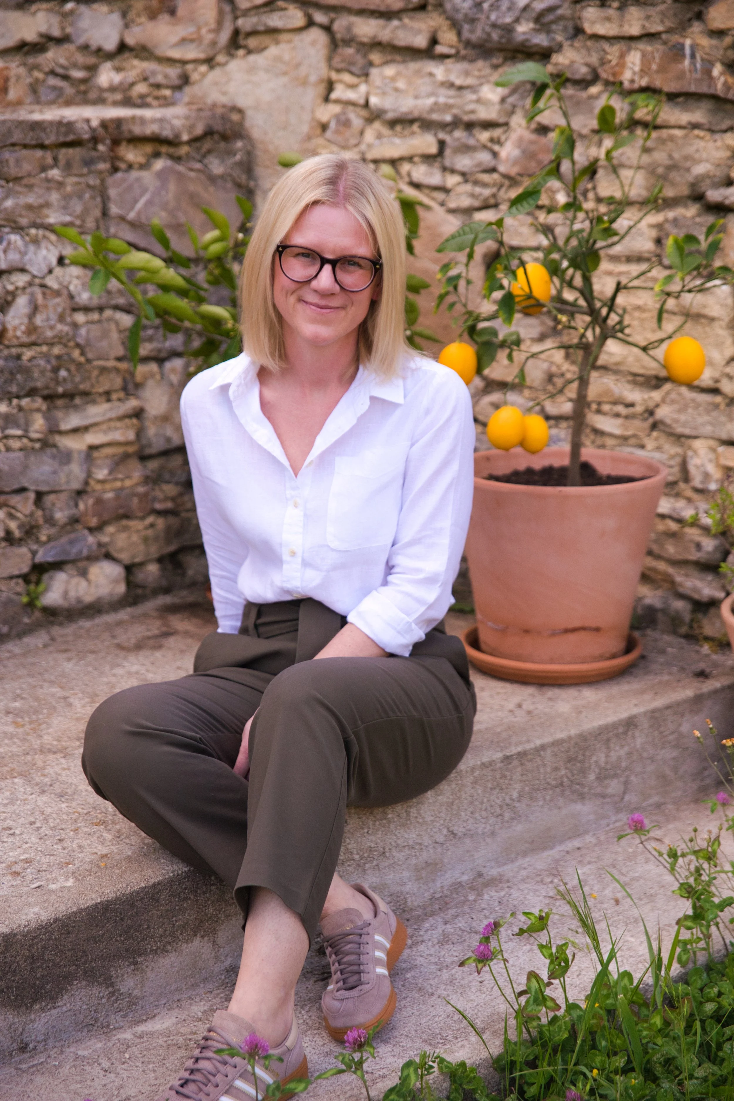 A woman with blonde hair, glasses, and a white shirt sitting on outdoor steps next to a potted lemon tree with yellow lemons in a garden with stone walls.