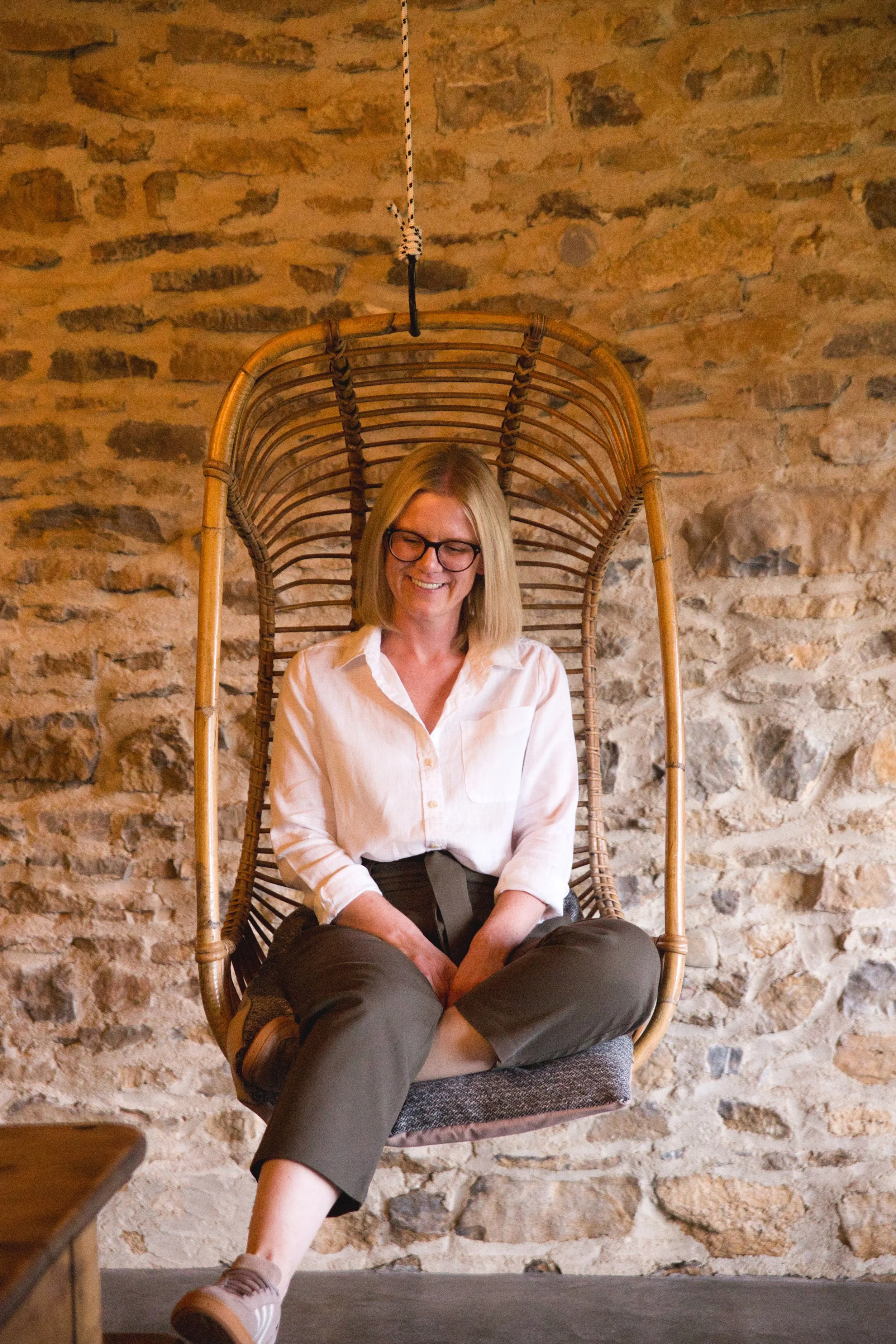 A woman with blonde hair and glasses sitting on a hanging rattan chair with a stone wall background, wearing a white shirt and gray pants.
