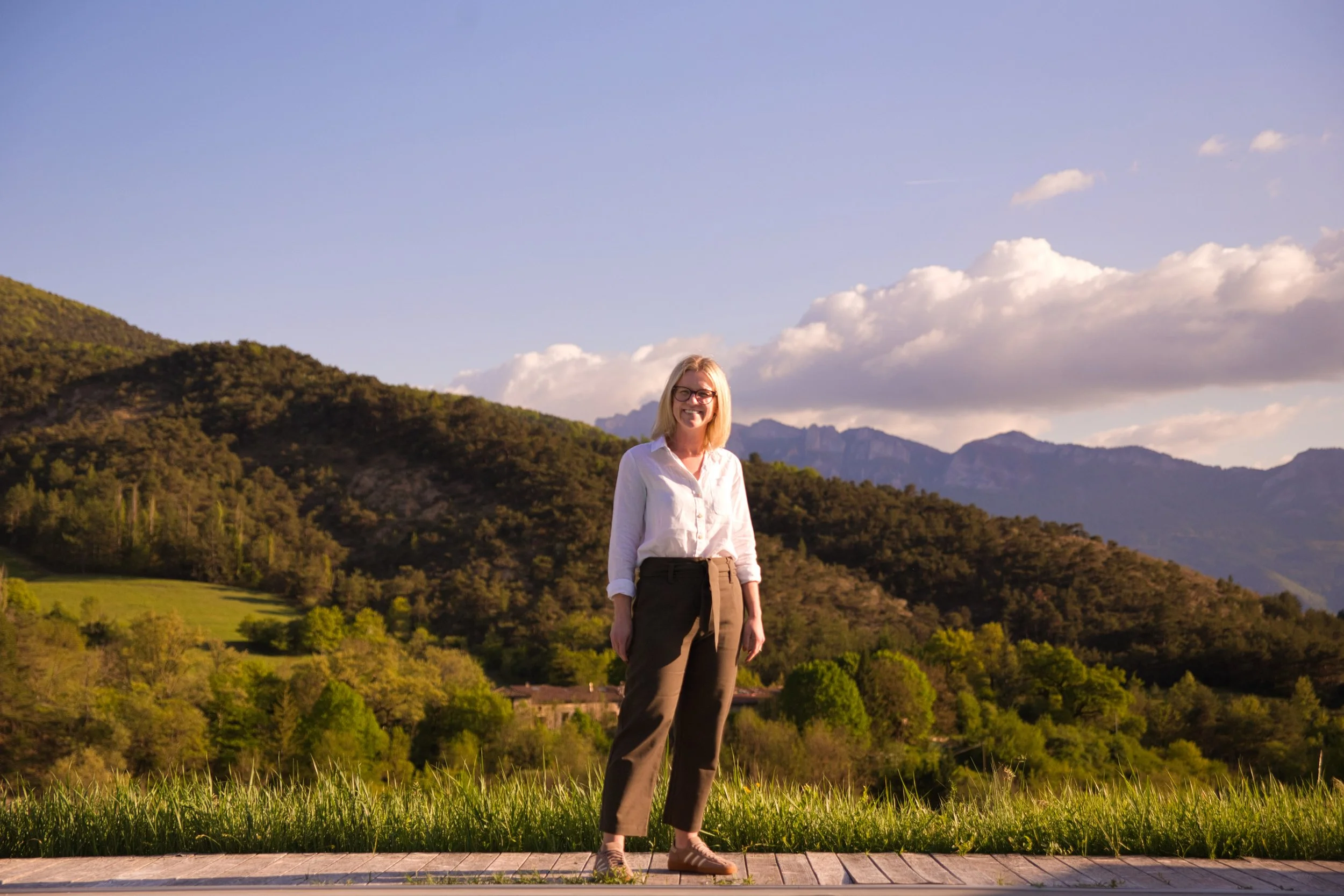A woman standing outdoors in front of green hills and mountains during sunset, wearing glasses, a light-colored blouse, and dark pants.