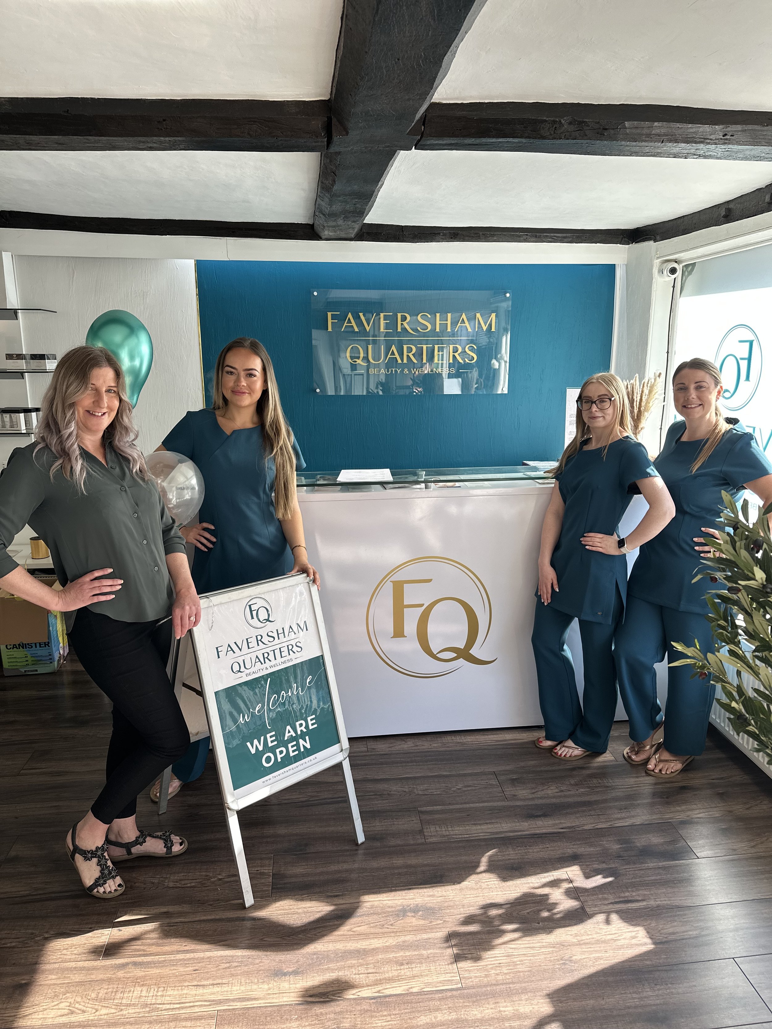 Four women standing behind a reception counter at Faversham Quarters Beauty and Wellness, with a sign in front that says the establishment is open. They are smiling and dressed in blue or gray uniforms.