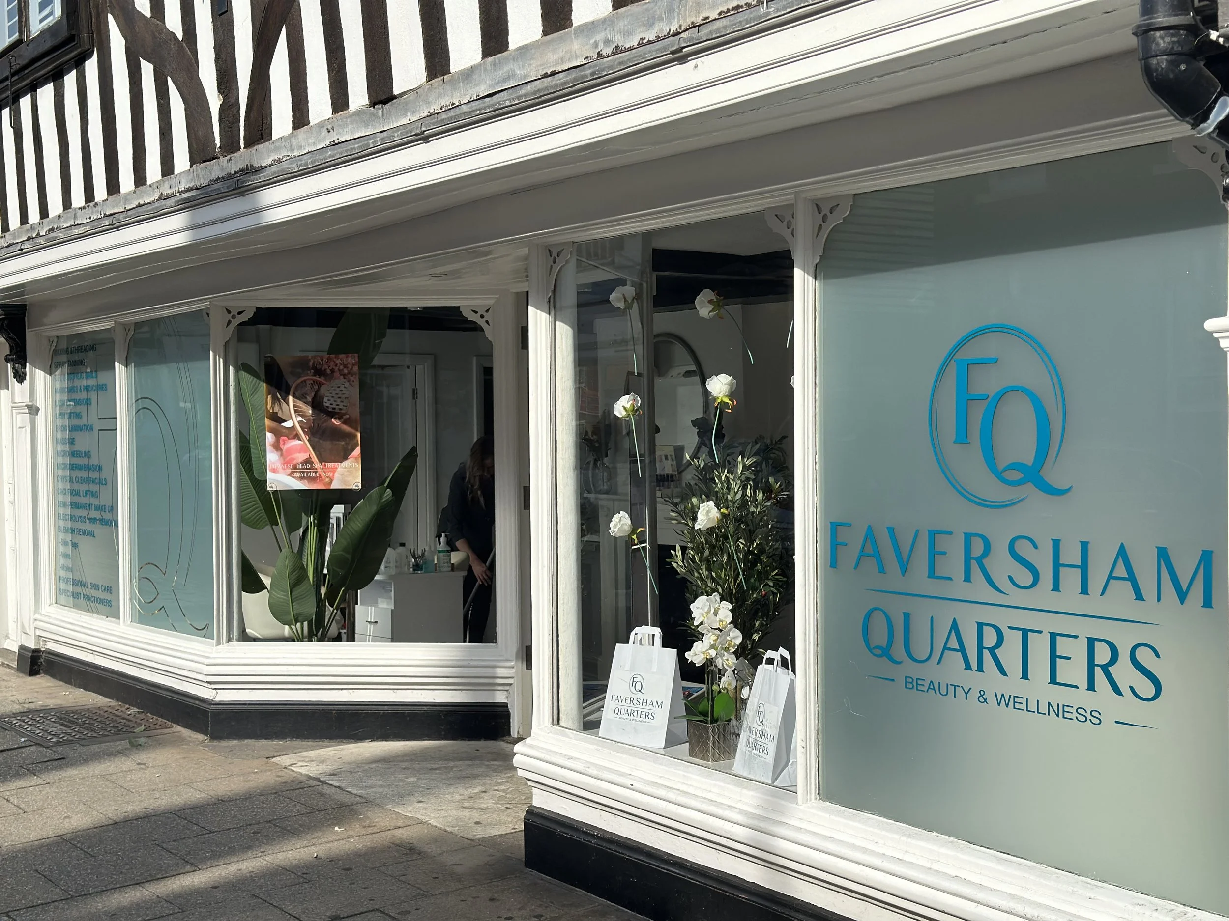 The image shows the exterior of Faversham Quarters, a beauty and wellness clinic. The storefront features large glass windows with white frames and decorative trim. Inside, there are white orchids and potted plants. There is a person visible inside, and signage displays the clinic's name and services.