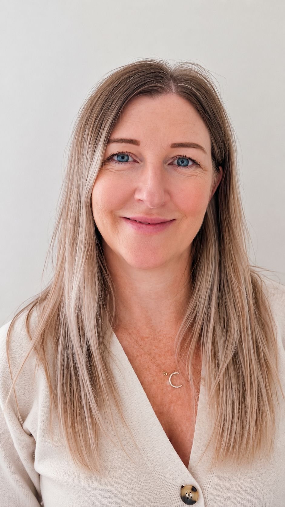 Portrait of a smiling woman with long blonde hair, blue eyes, wearing a beige coat with large buttons, and a gold necklace with a 'C' pendant, standing against a plain white background.