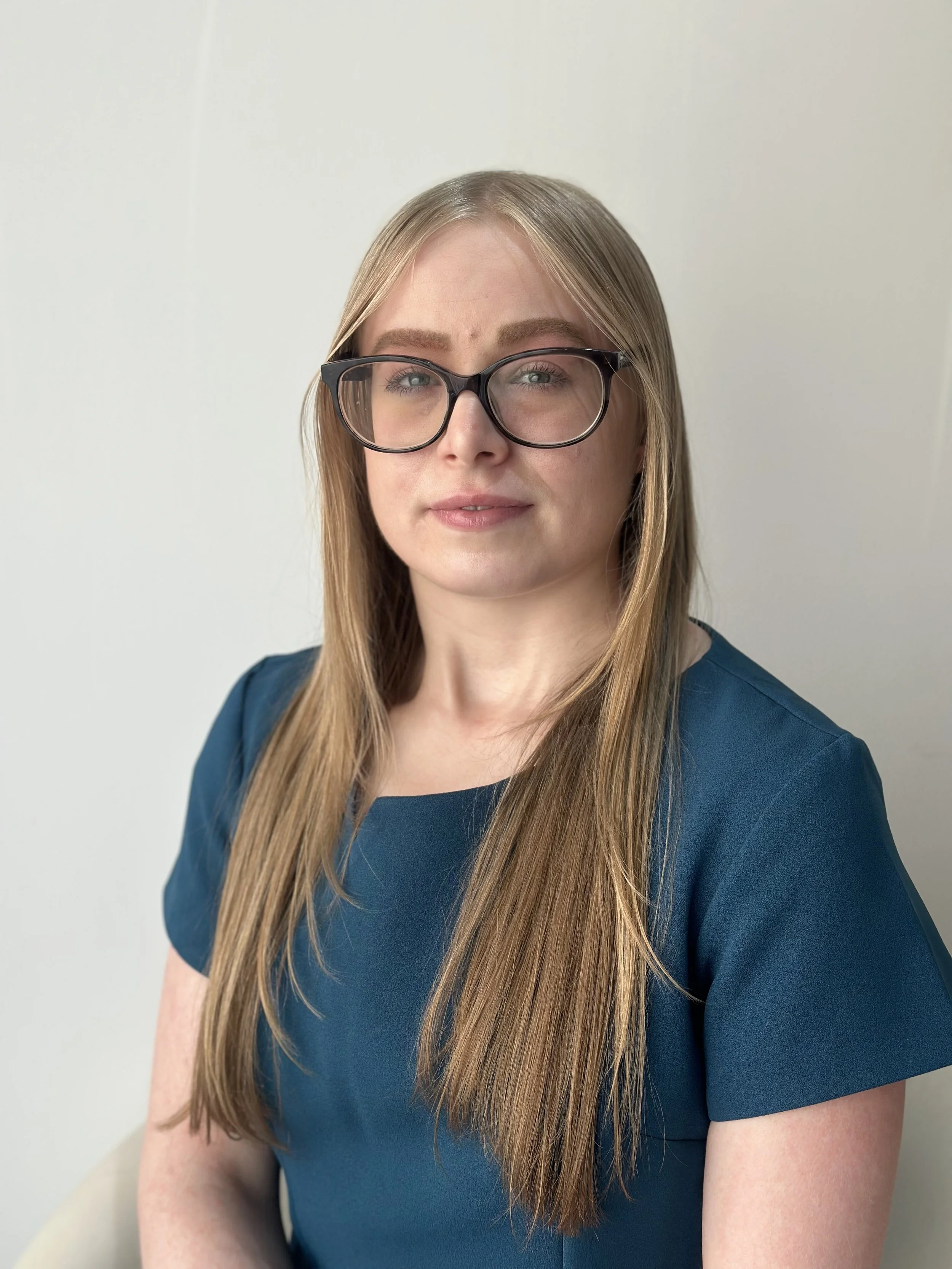 A woman with long, straight, light brown hair wearing glasses and a dark teal top, sitting against a plain white wall.