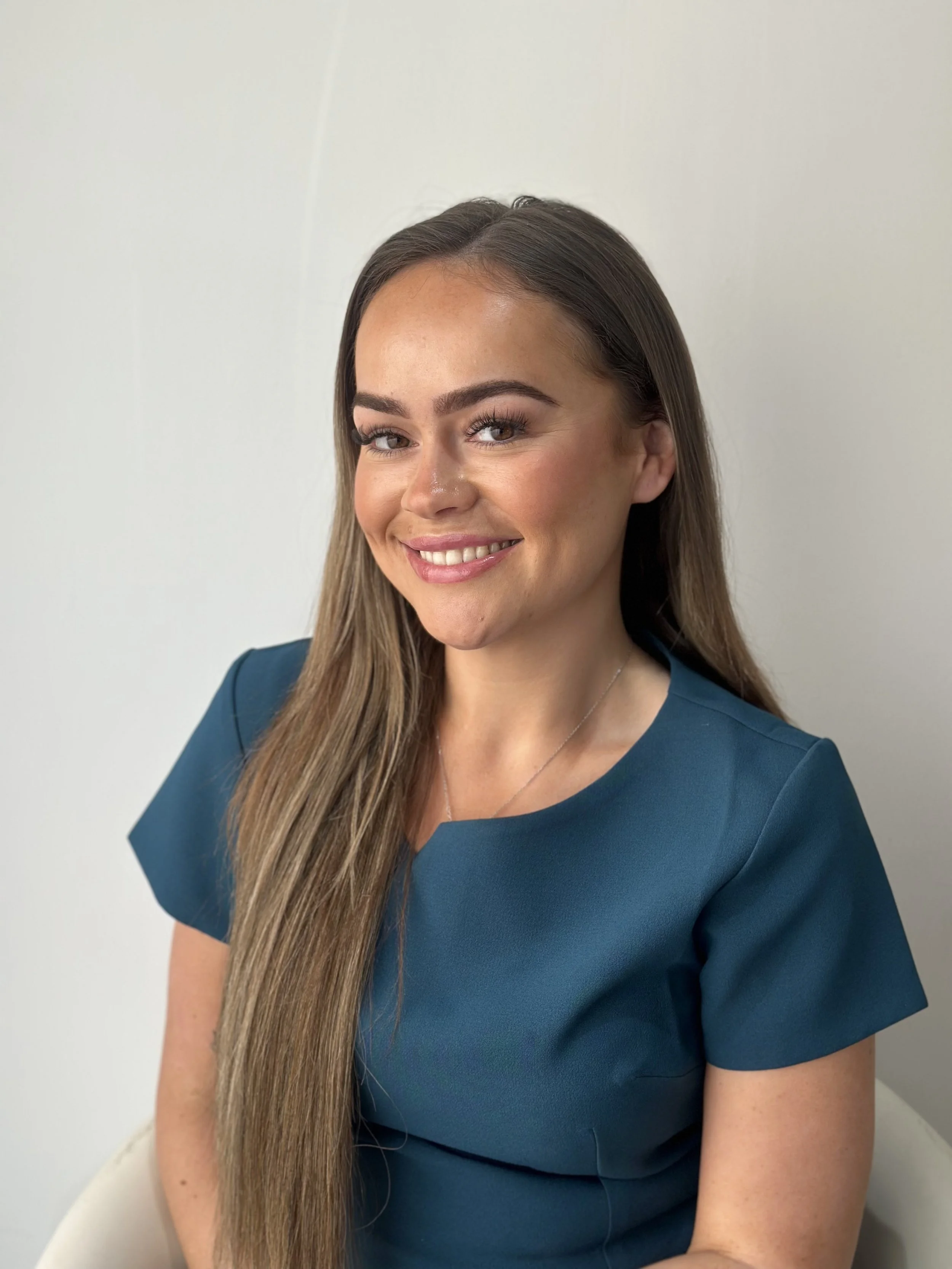 A young woman with long brown hair, smiling, wearing a dark teal top, against a plain light background.