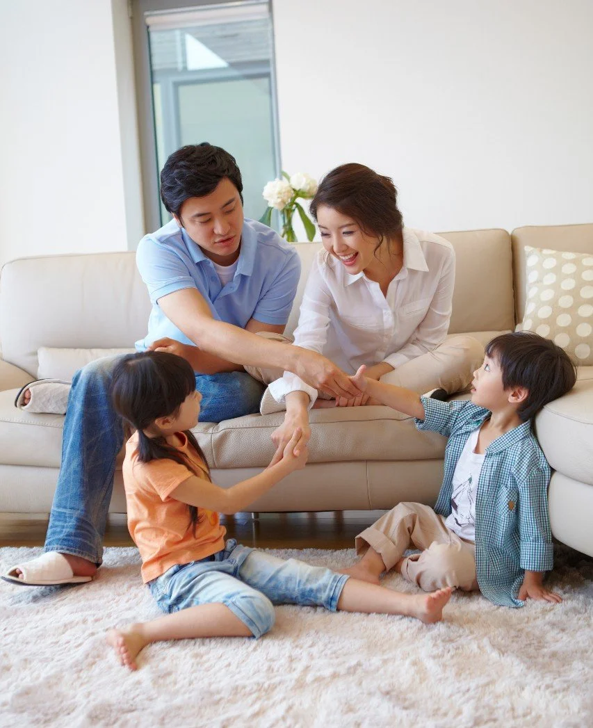 A family of four sitting on a beige carpet in their living room. The parents are sitting on a couch, smiling and engaging with their two children who are sitting on the floor. The mother is reaching out to touch the daughter's hand, while the father is reaching toward the son's hand. The children are facing each other, holding hands, and appear to be playing or sharing a moment of affection.