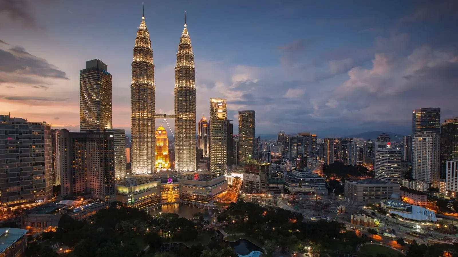 Nighttime city skyline featuring the illuminated Petronas Towers in Kuala Lumpur, Malaysia, surrounded by other tall buildings with lights.
