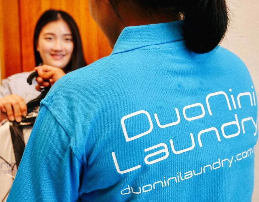Person wearing a blue polo shirt with 'Duonini Laundry' logo, facing away, with a woman in the background holding a laundry basket.