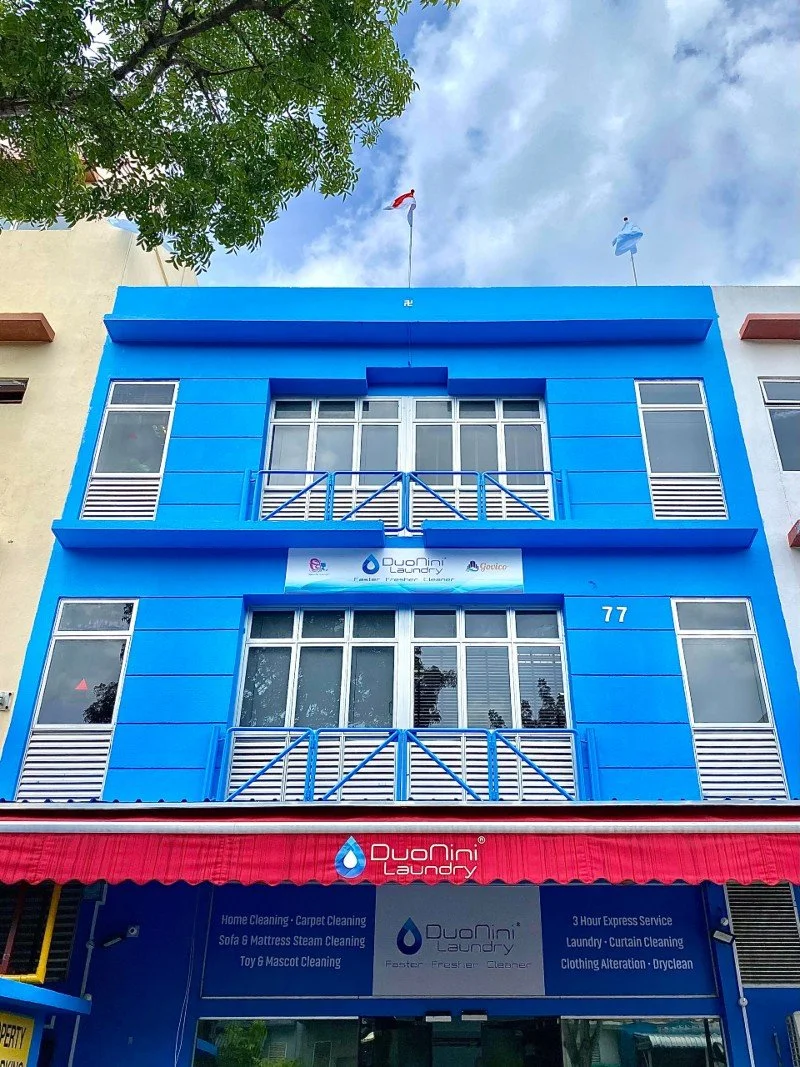 Photo of a three-story building painted bright blue, with a red and a blue flag on top, and a sign for DuoNini Laundry on the ground floor.