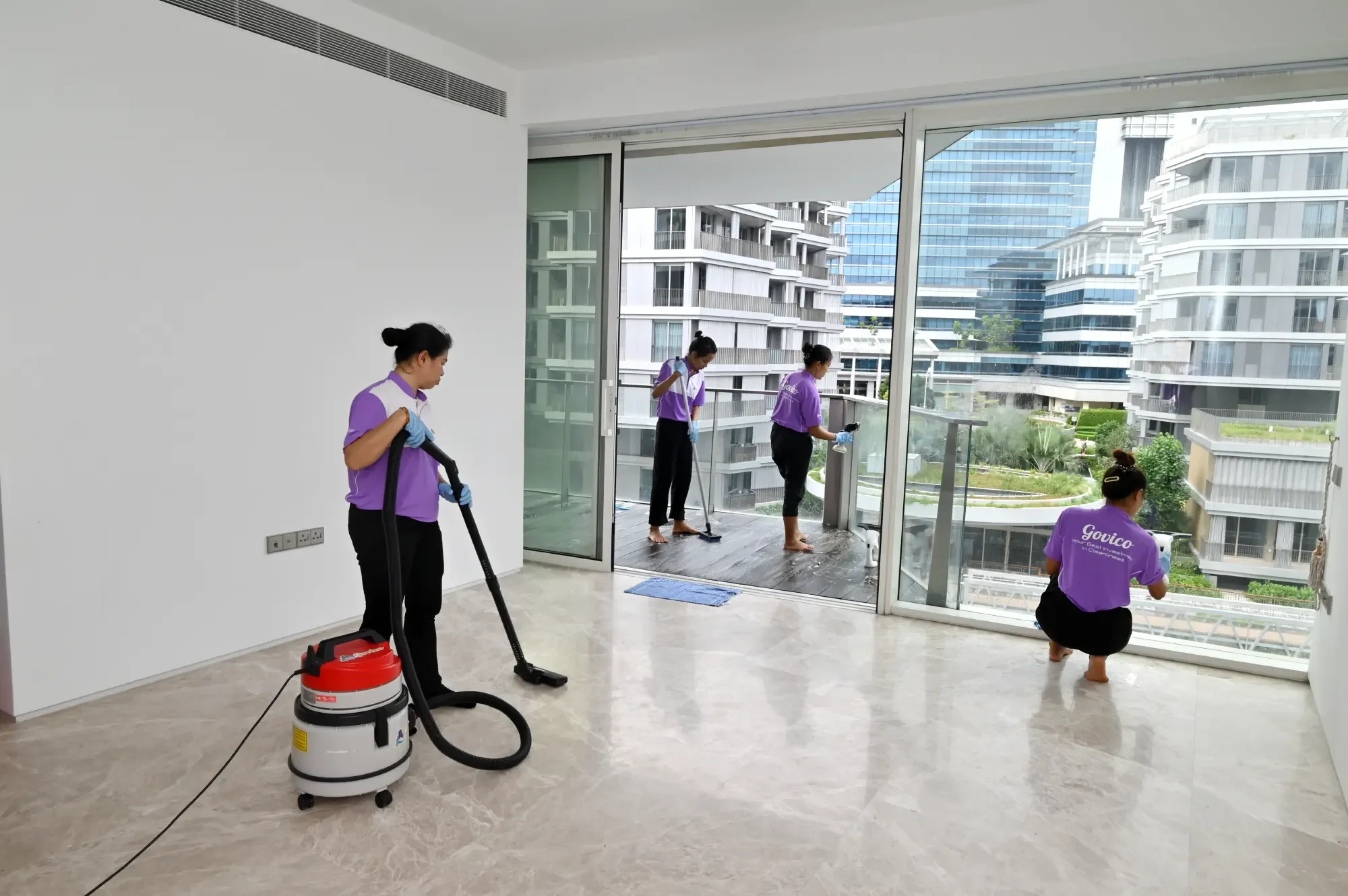 Five cleaning workers in purple uniforms cleaning a large apartment window, with a cityscape of modern buildings outside.