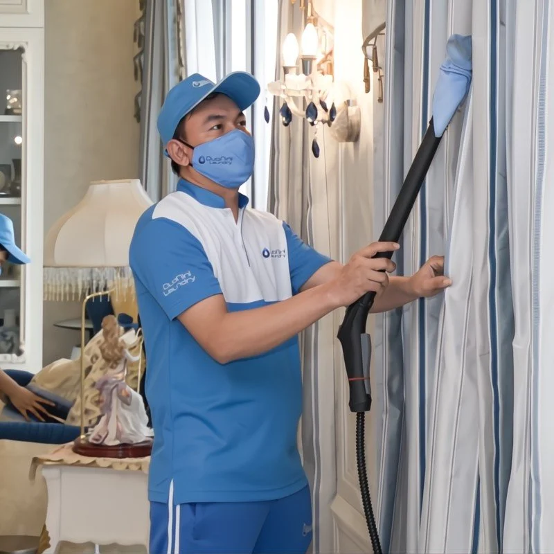 A person wearing a blue uniform and a face mask is vacuuming curtains in a room.