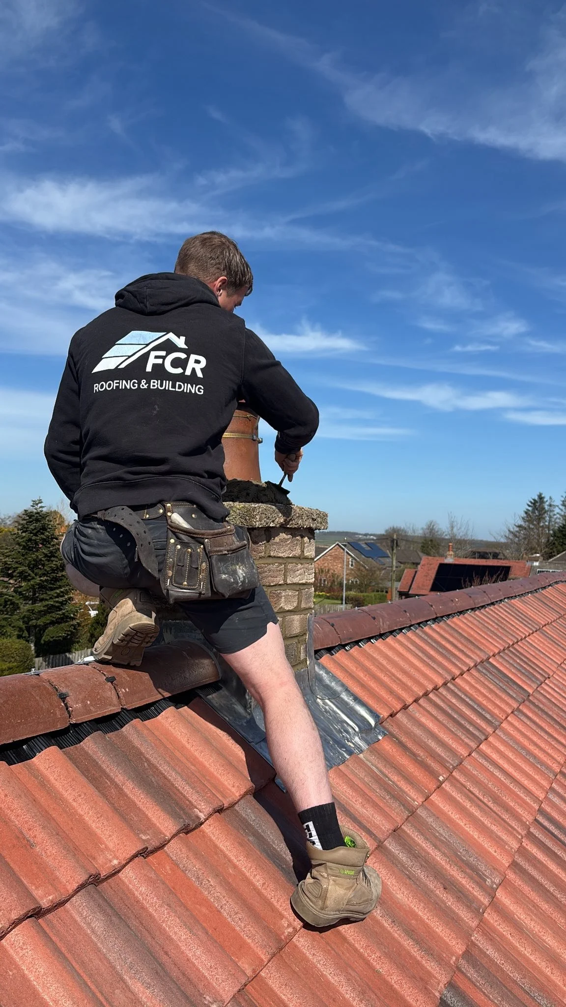 A man working on a red tile roof, wearing a black hoodie with a logo, and gear; he is positioned on the roof near a chimney, with a neighborhood and blue sky in the background.