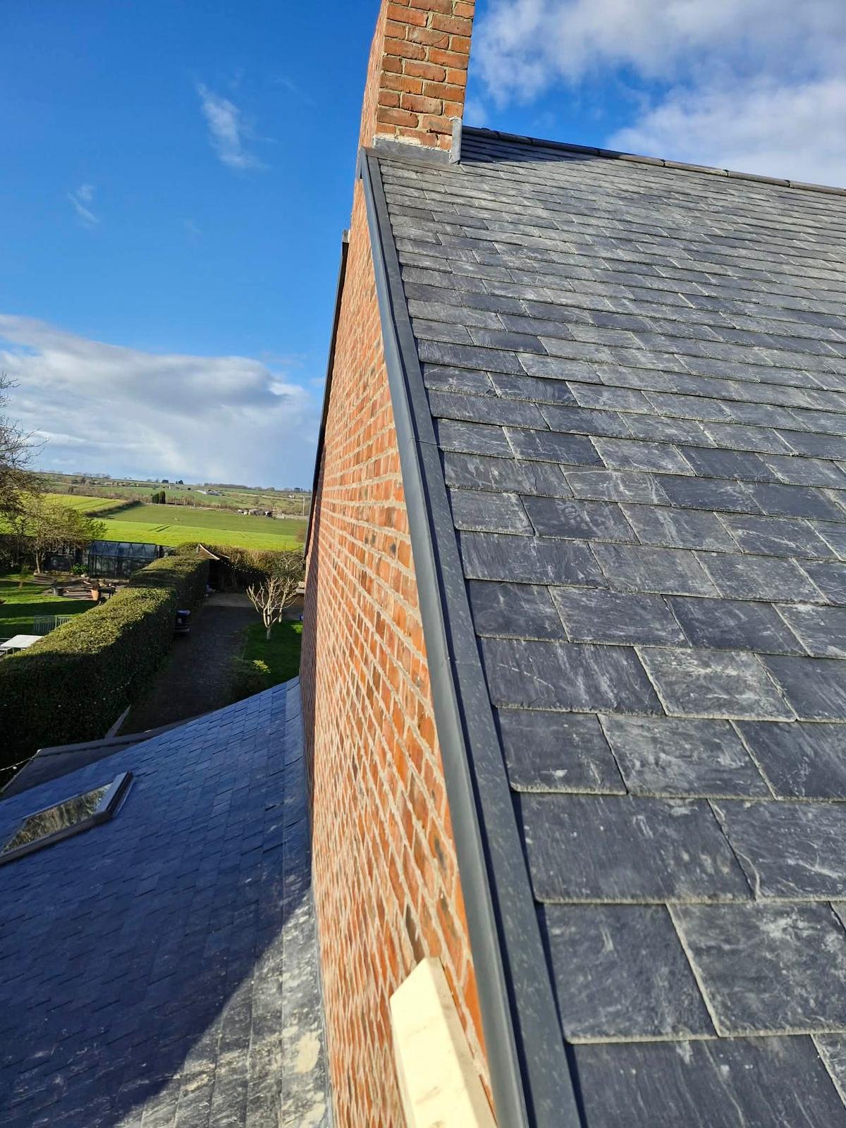 A view of a roof with slate tiles, a brick chimney, and a brick wall under a blue sky with clouds in a rural landscape.