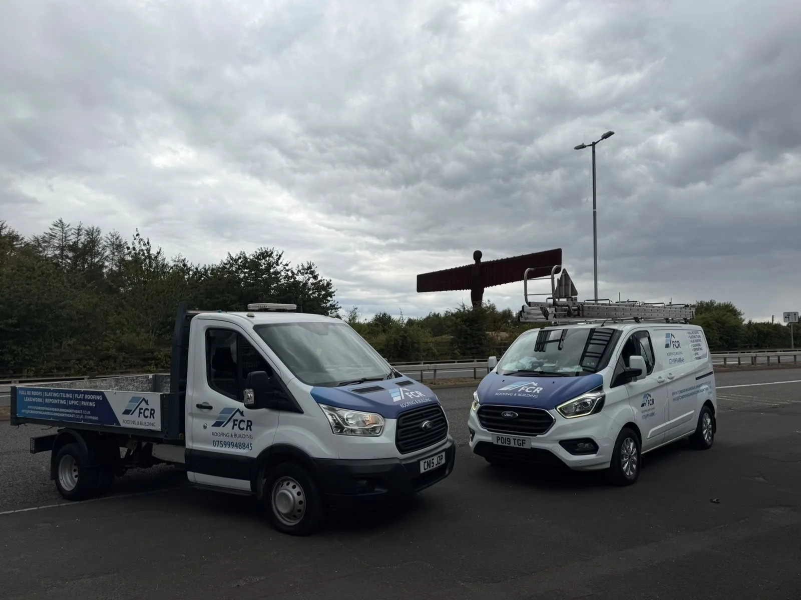 Two FCR roofing and building service trucks parked on the side of a road under a cloudy sky, with trees and road signs in the background.