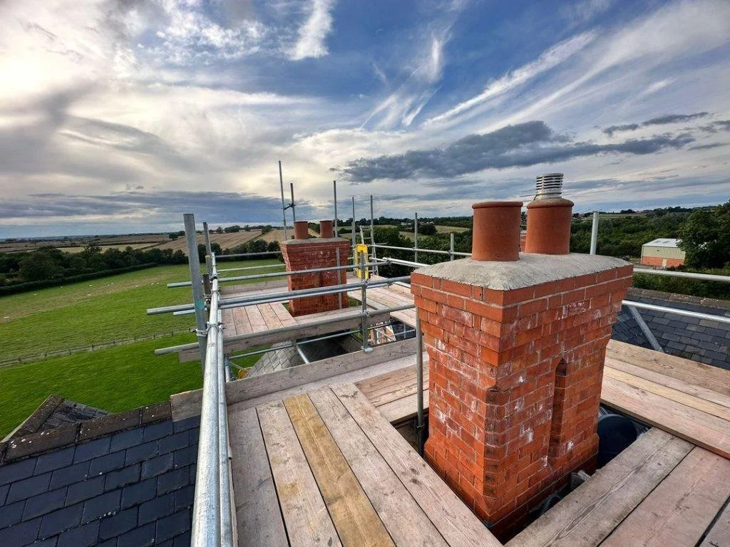 View of a house roof with brick chimneys, surrounded by scaffolding, overlooking a green field and distant trees under a partly cloudy sky.